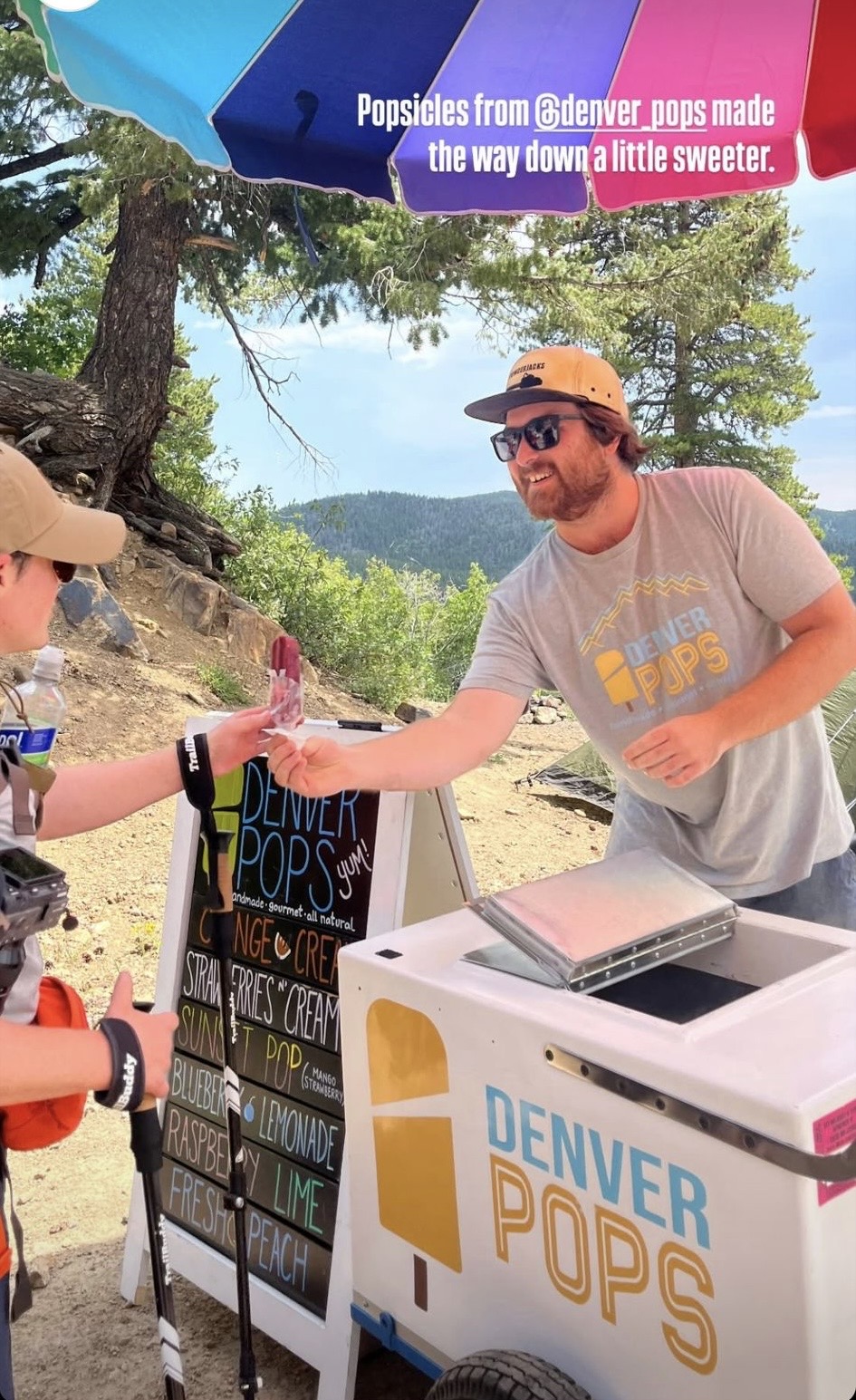 Person handing money to vendor at Denver Pops ice cream stand outdoors, with trees and a hill in background.
