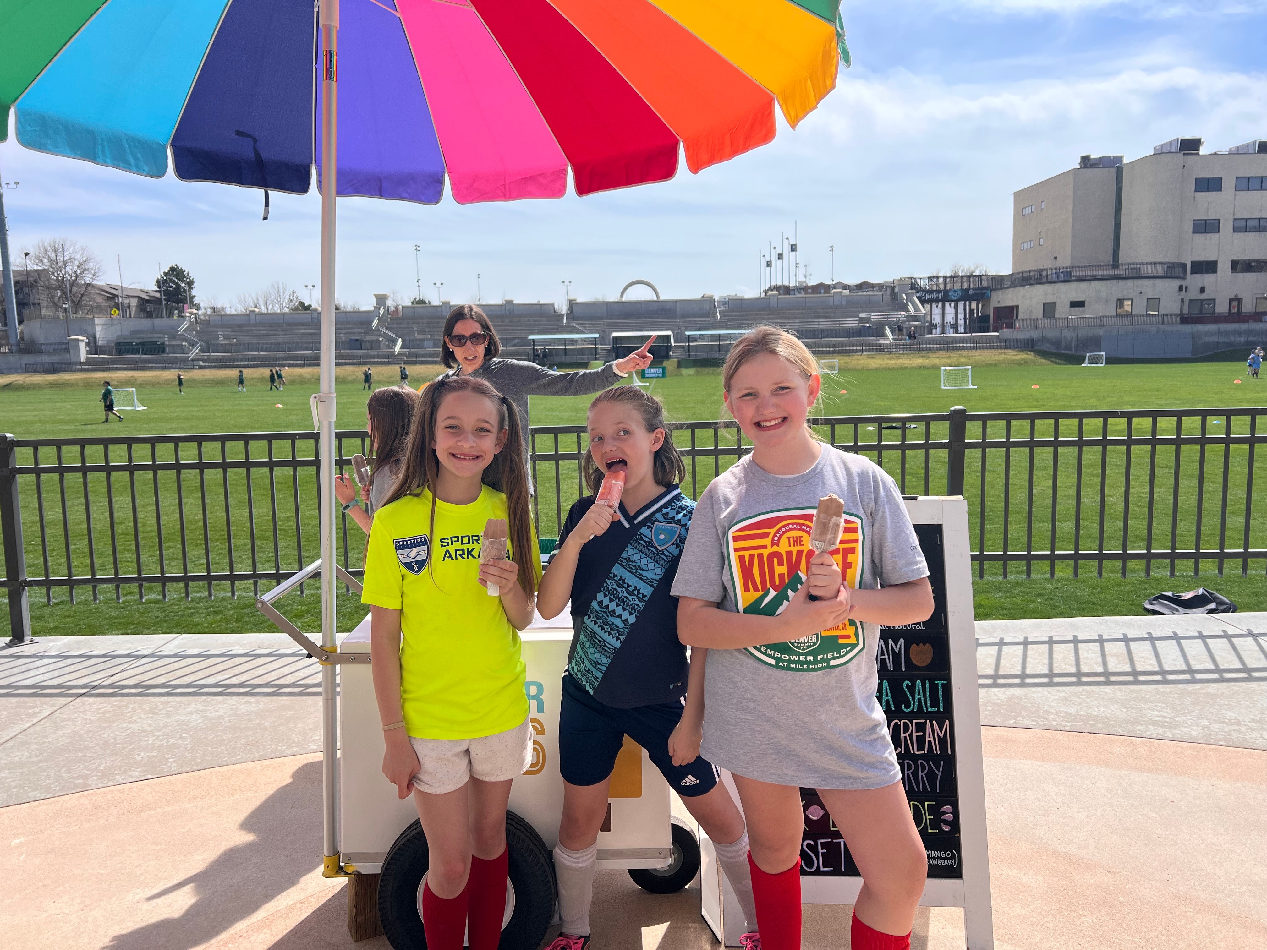 Four children stand outdoors near a colorful umbrella, with a grassy field and buildings in the background.