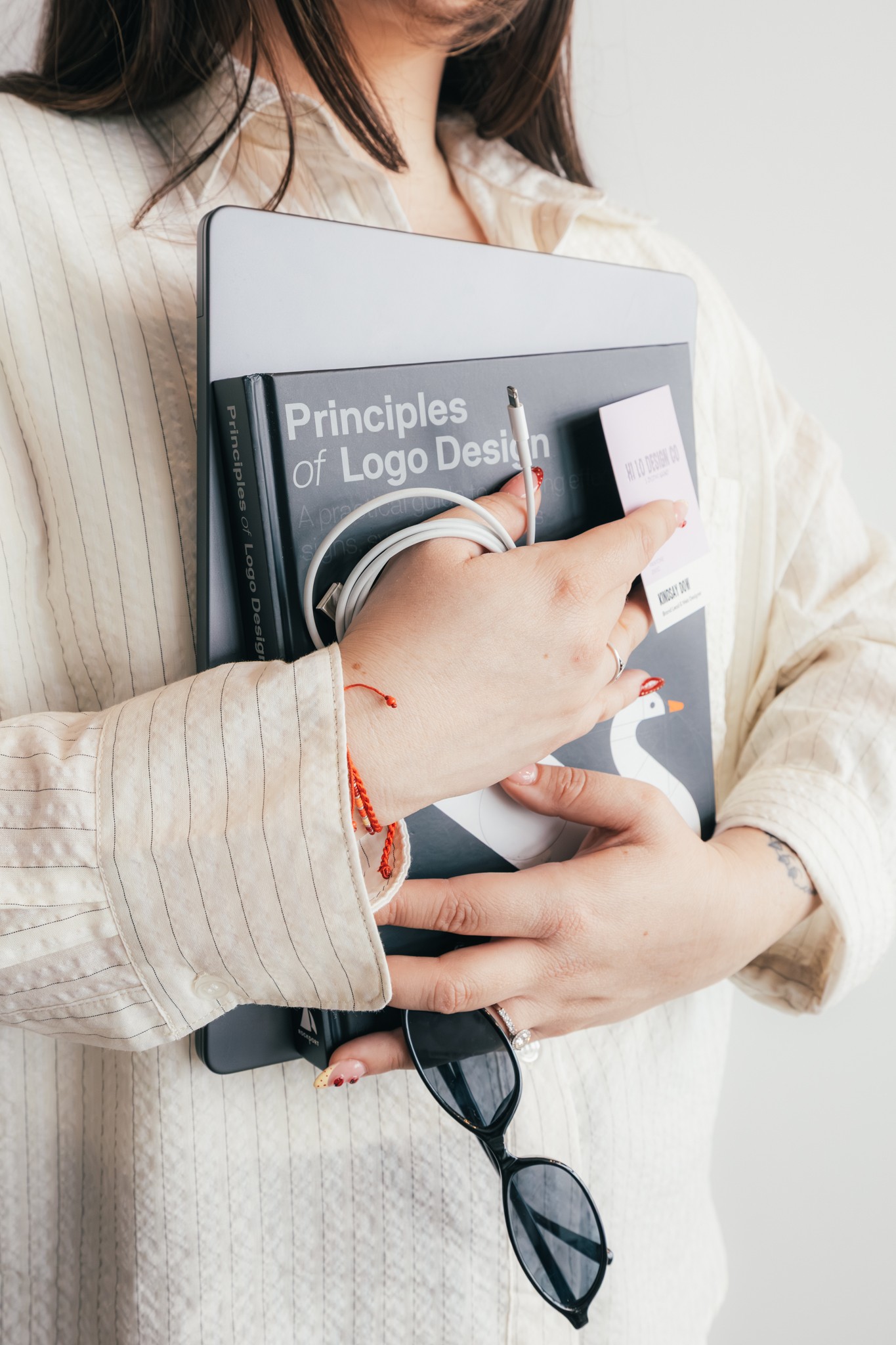 Person holding a book titled 'Principles of Logo Design' and a smartphone, wearing a beige striped shirt, with sunglasses hanging from the book.