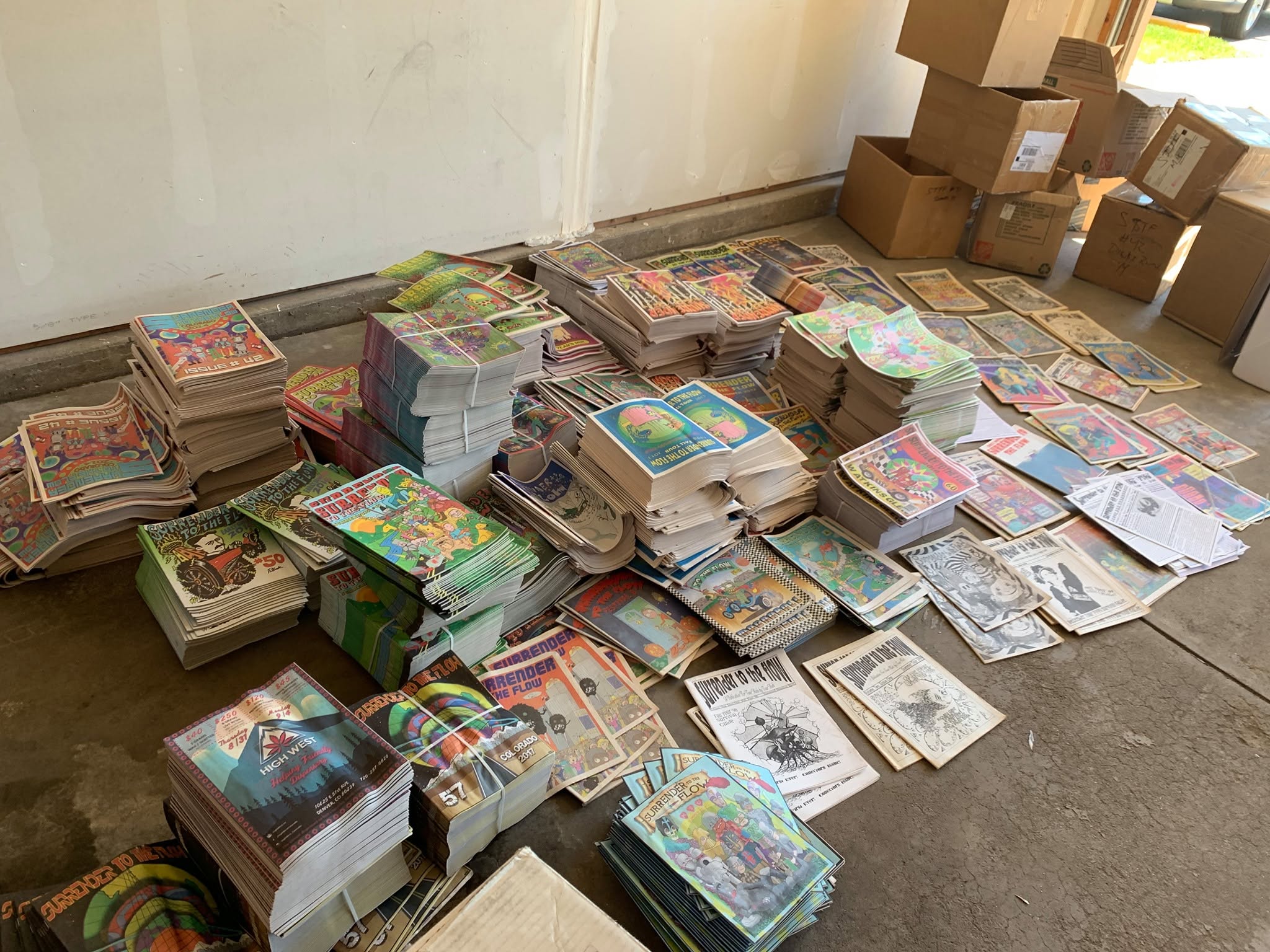 Stacks of colorful magazines and newspapers spread on the floor near cardboard boxes, with a plain wall in the background.