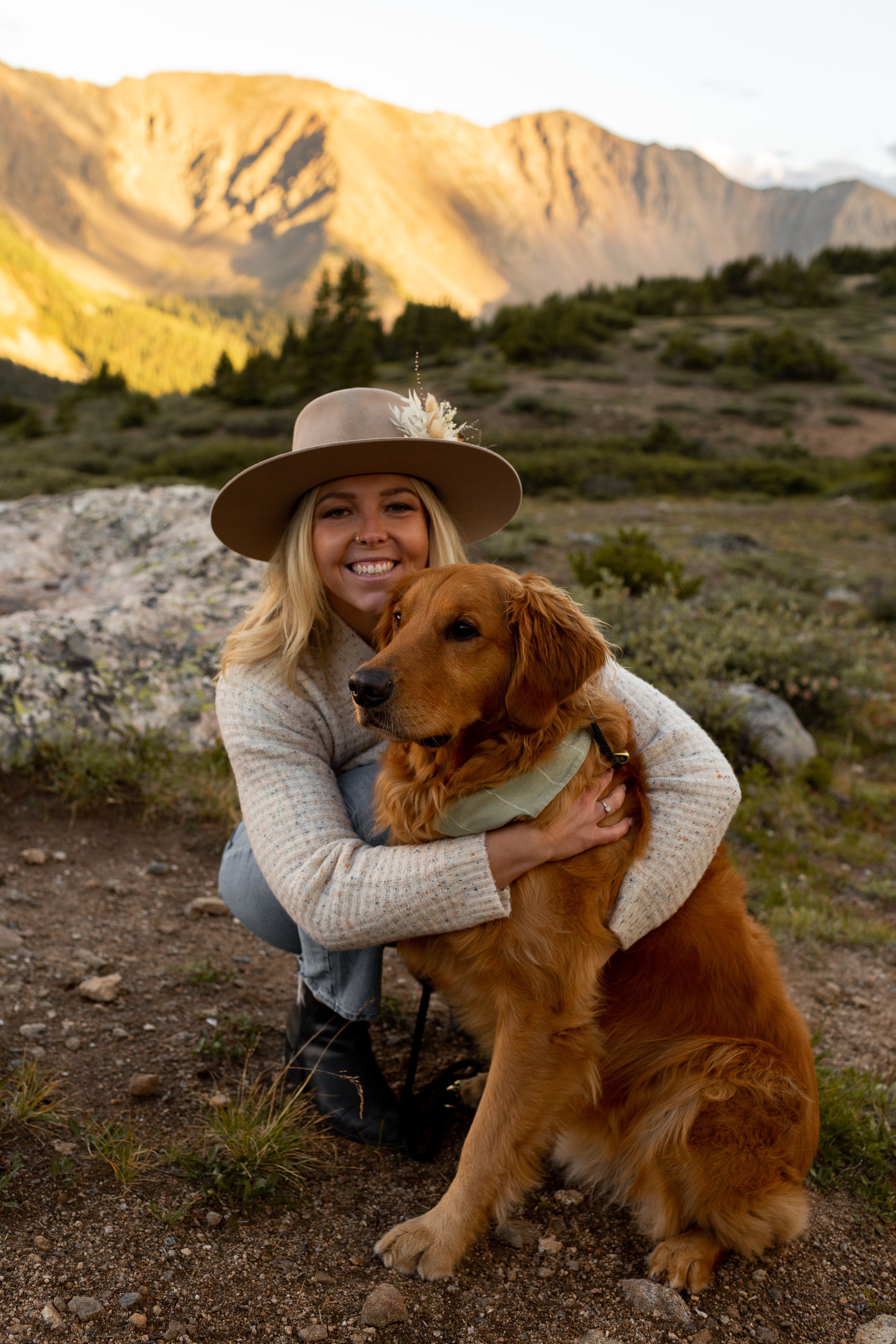Woman smiling with a golden retriever in a mountainous landscape, wearing a hat and sweater.