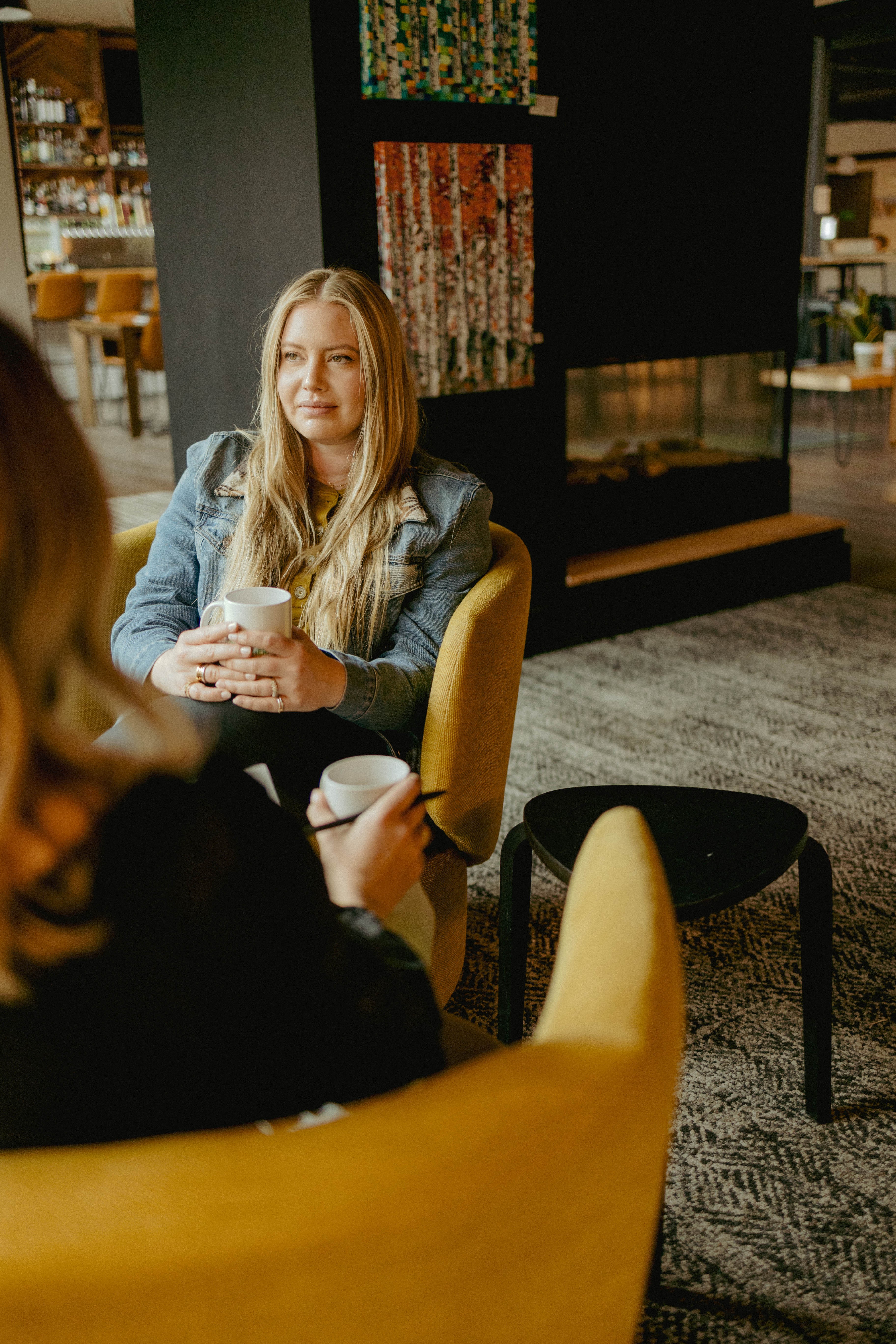 Young woman with long blonde hair sitting in a yellow chair, holding a cup, in a modern indoor setting.