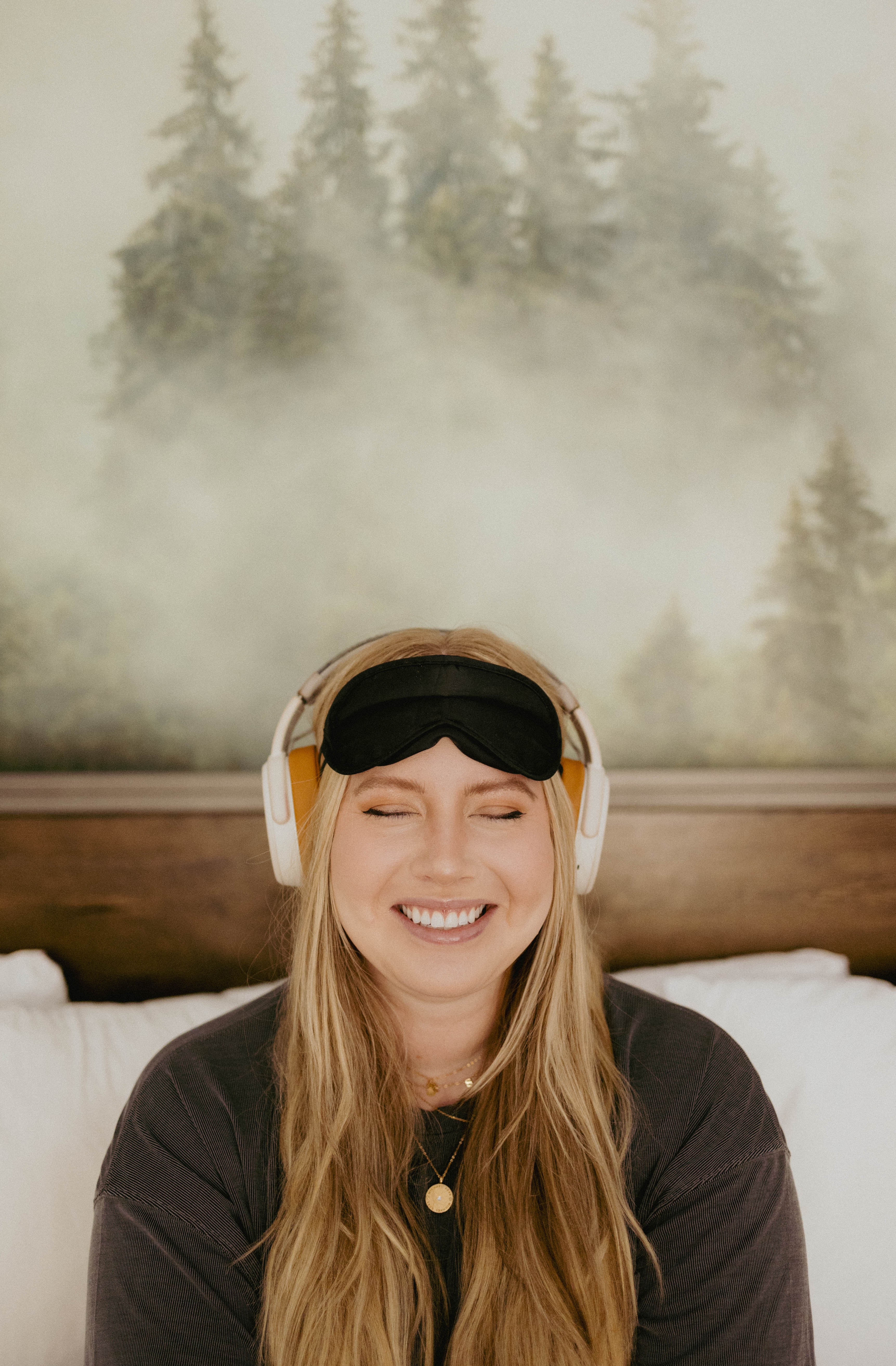 Young woman with long hair wearing headphones and a sleep mask, smiling with eyes closed, in a bedroom with forest mural.