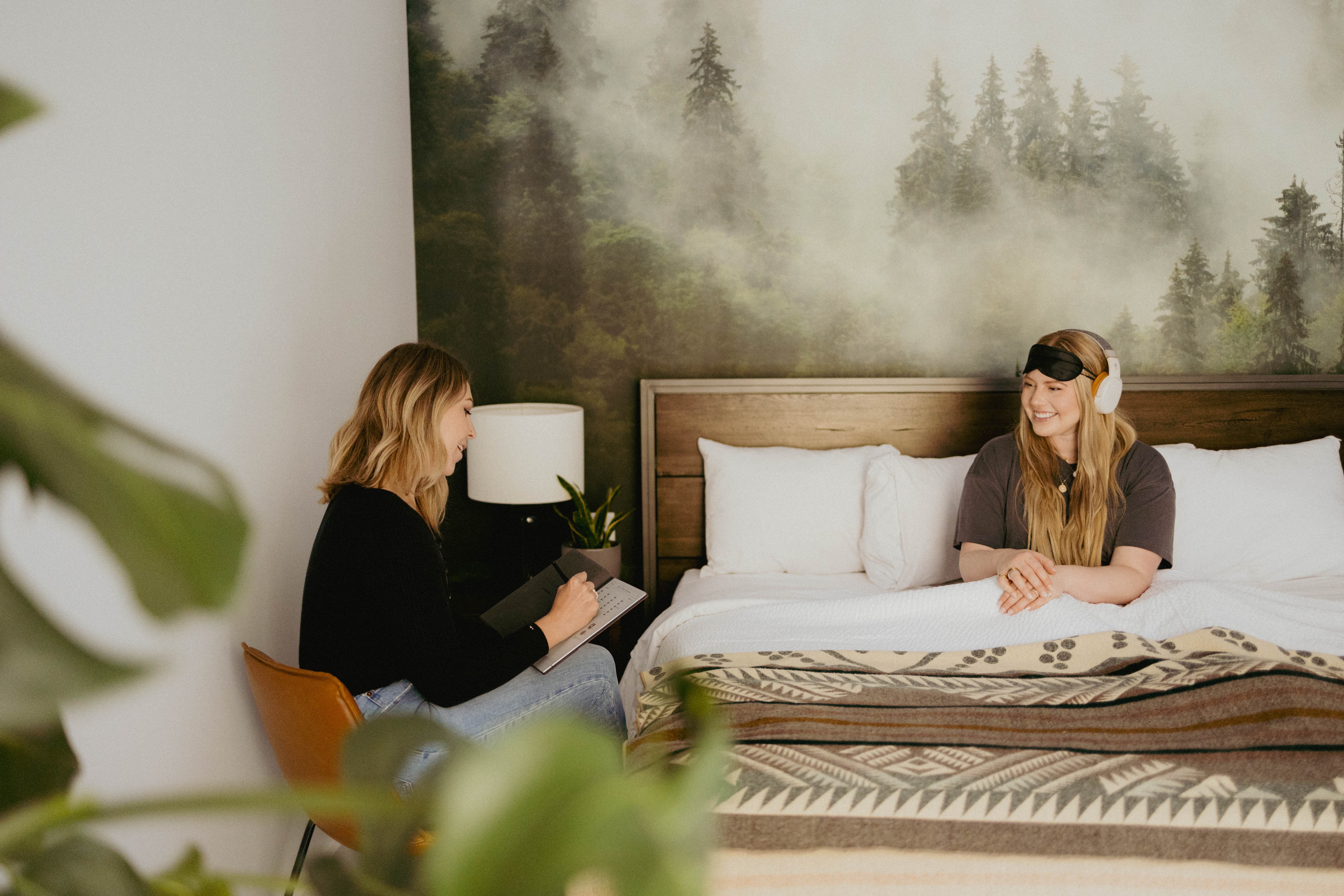 Two women sitting on a bed in a room with a forest mural, one writing and the other smiling, with a nightstand and lamp nearby.