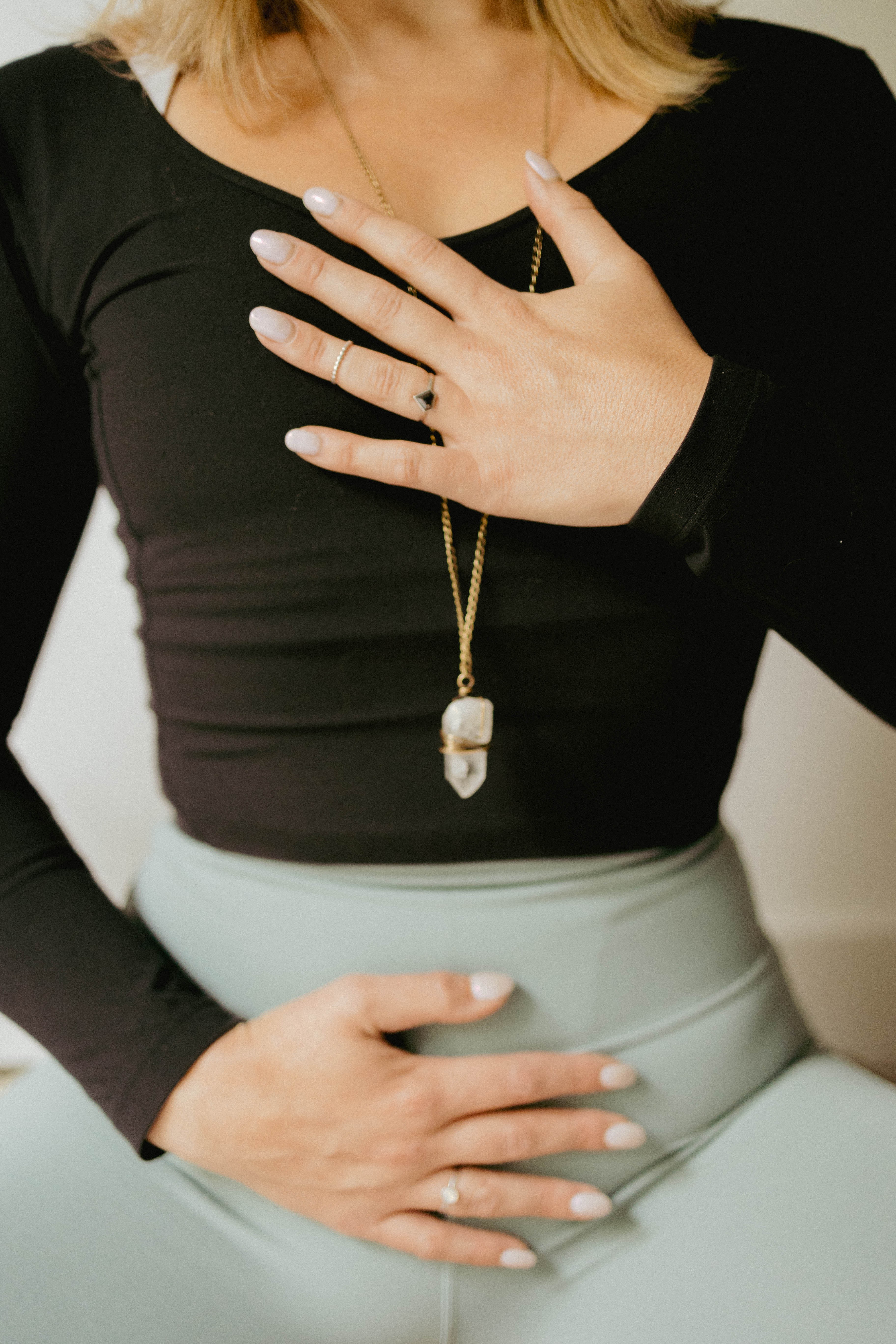 Person wearing a black top and light-colored skirt, with hand on chest and another on abdomen, wearing a necklace with a pendant.