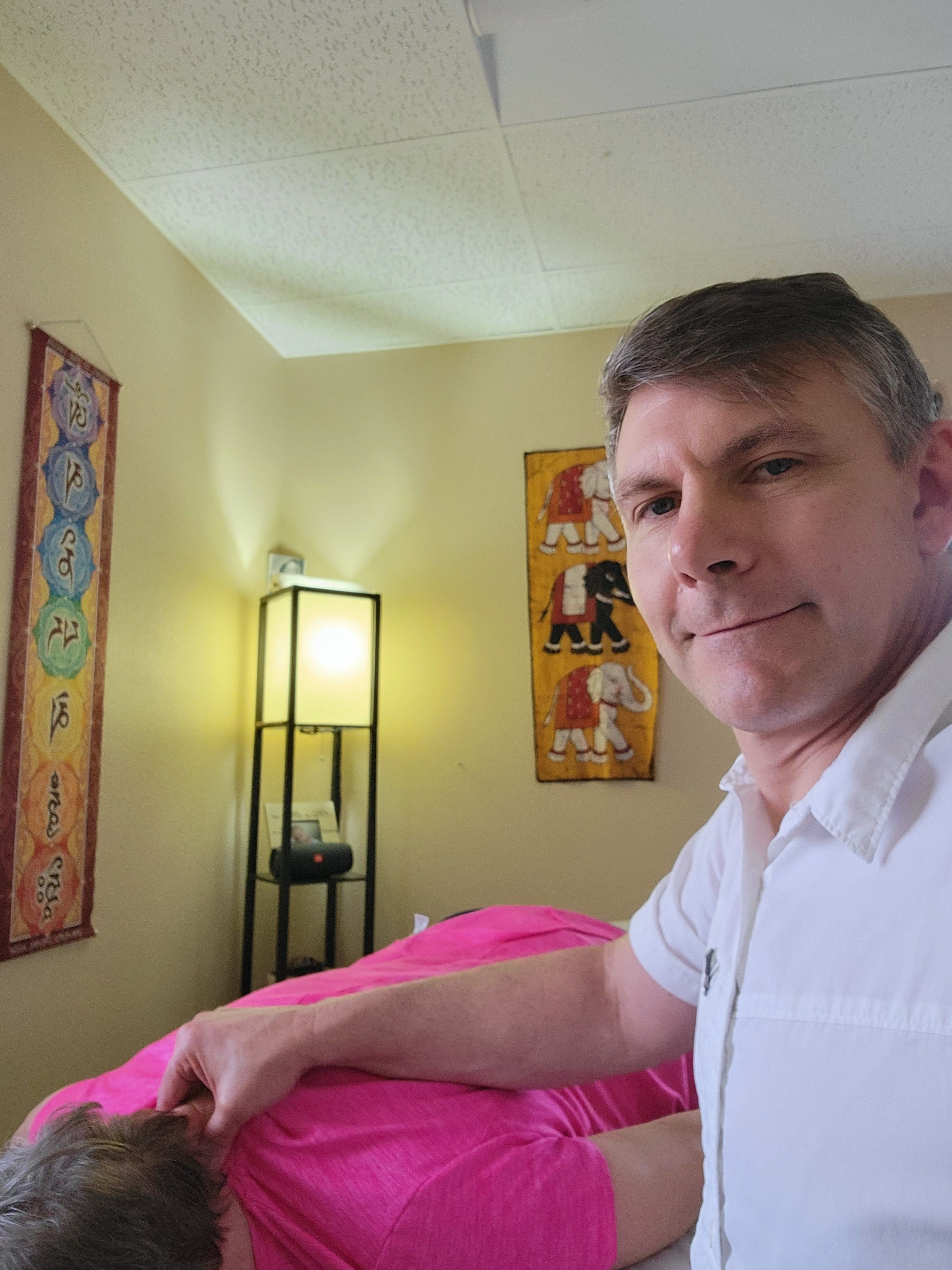 Person lying on a bed with pink bedding, in a room with colorful wall art and a lamp, facing the camera.
