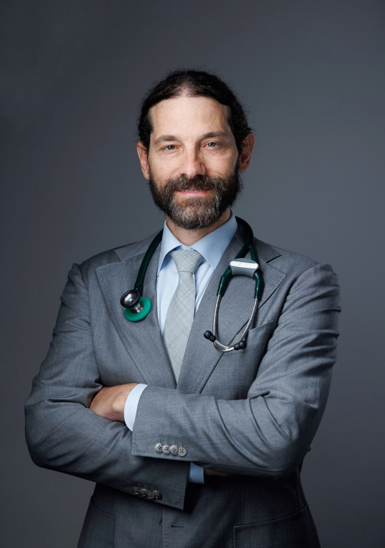 Man with a beard in a suit and stethoscope crosses arms, smiling, against a dark background.