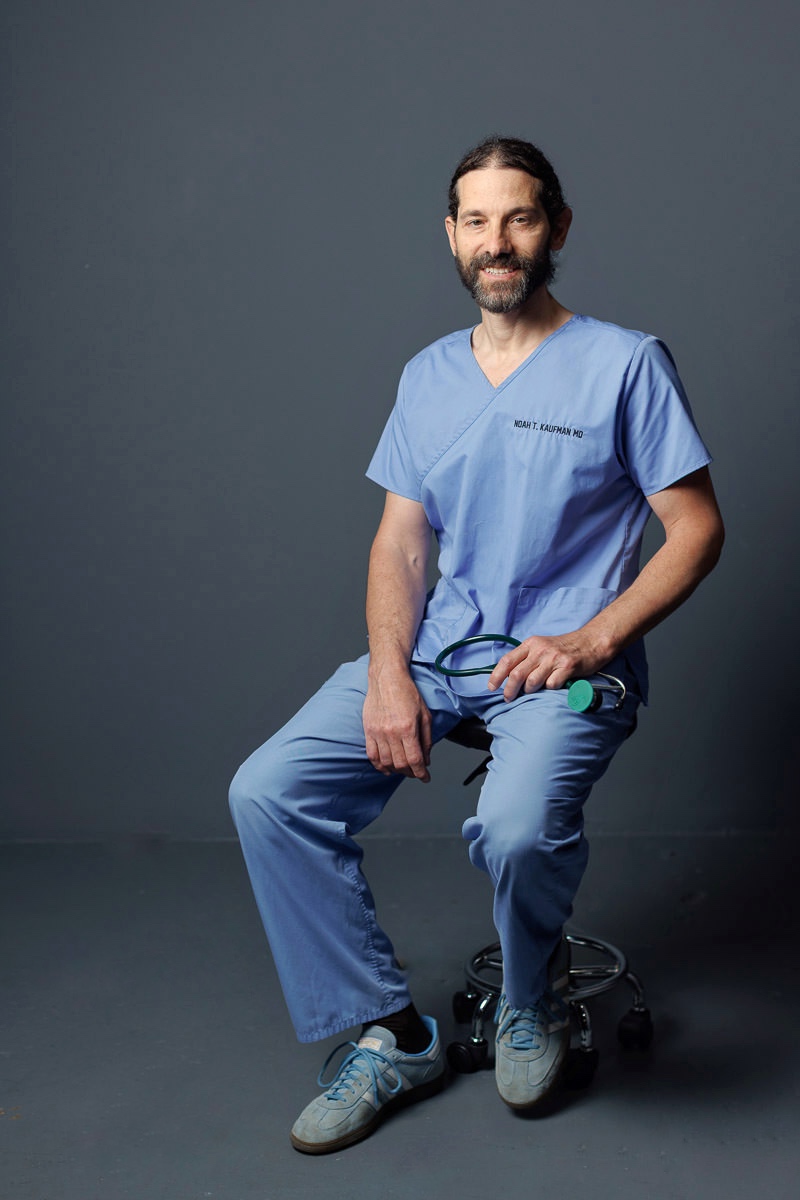 Man in blue medical scrubs sitting on a stool, smiling, with a plain dark background.