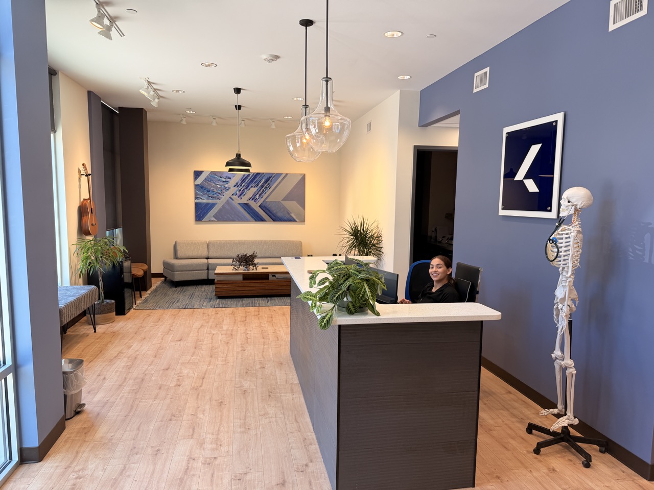 Reception area with a person sitting behind a counter, blue and beige walls, artwork, plants, and a skeleton model.