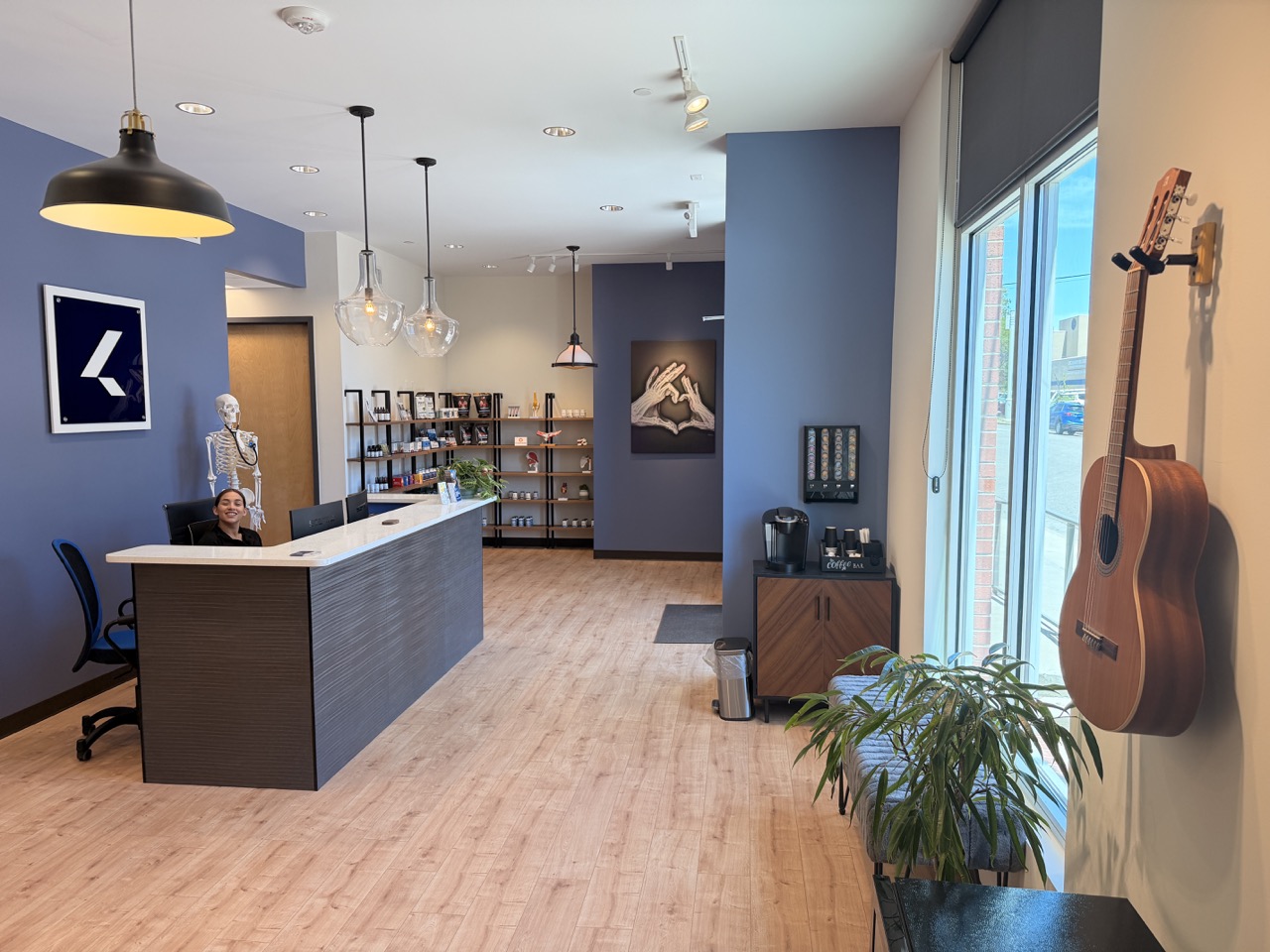 Reception area with a desk, bookshelf, and wall art, large window with a guitar hanging, and a plant near the window.