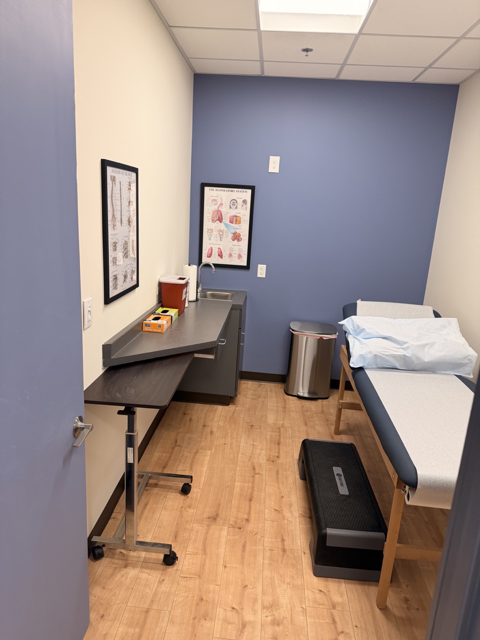Hospital examination room with bed, desk, framed posters, trash can, and wooden floor.