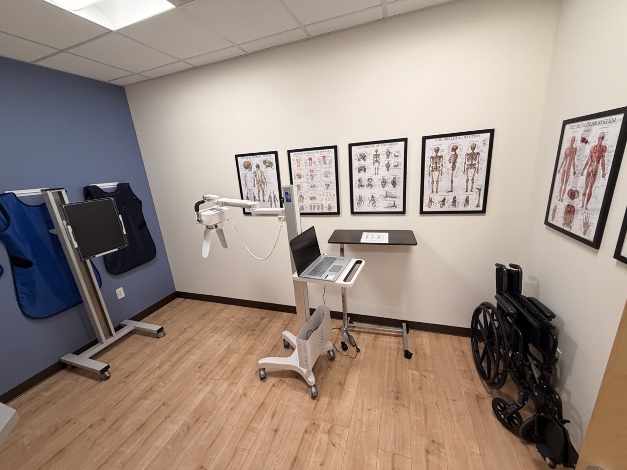 Medical equipment and anatomical posters on the wall in a clinical room with wooden flooring.
