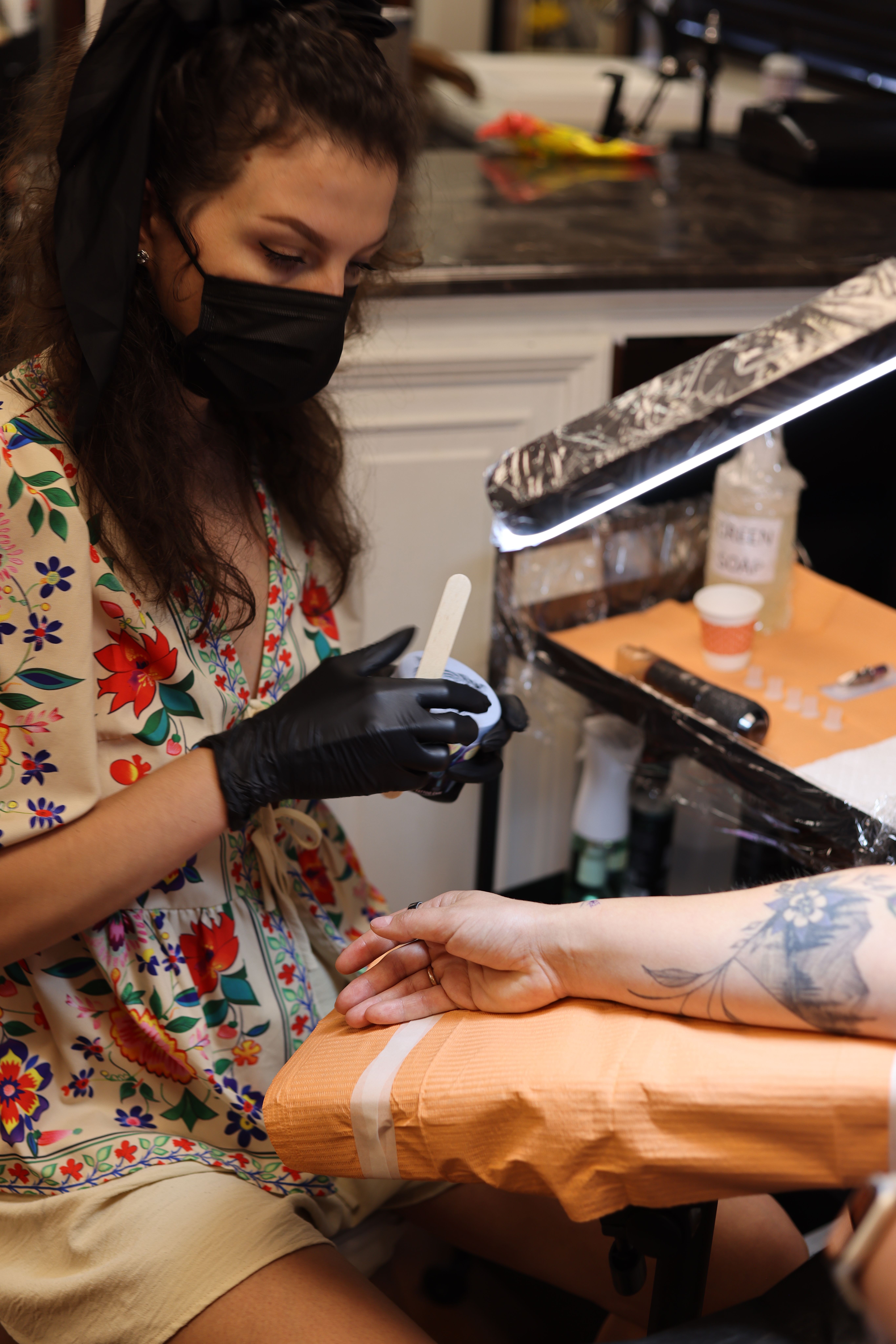 Person receiving a tattoo on their arm while a tattoo artist works, with tattoo equipment visible, in a tattoo studio.