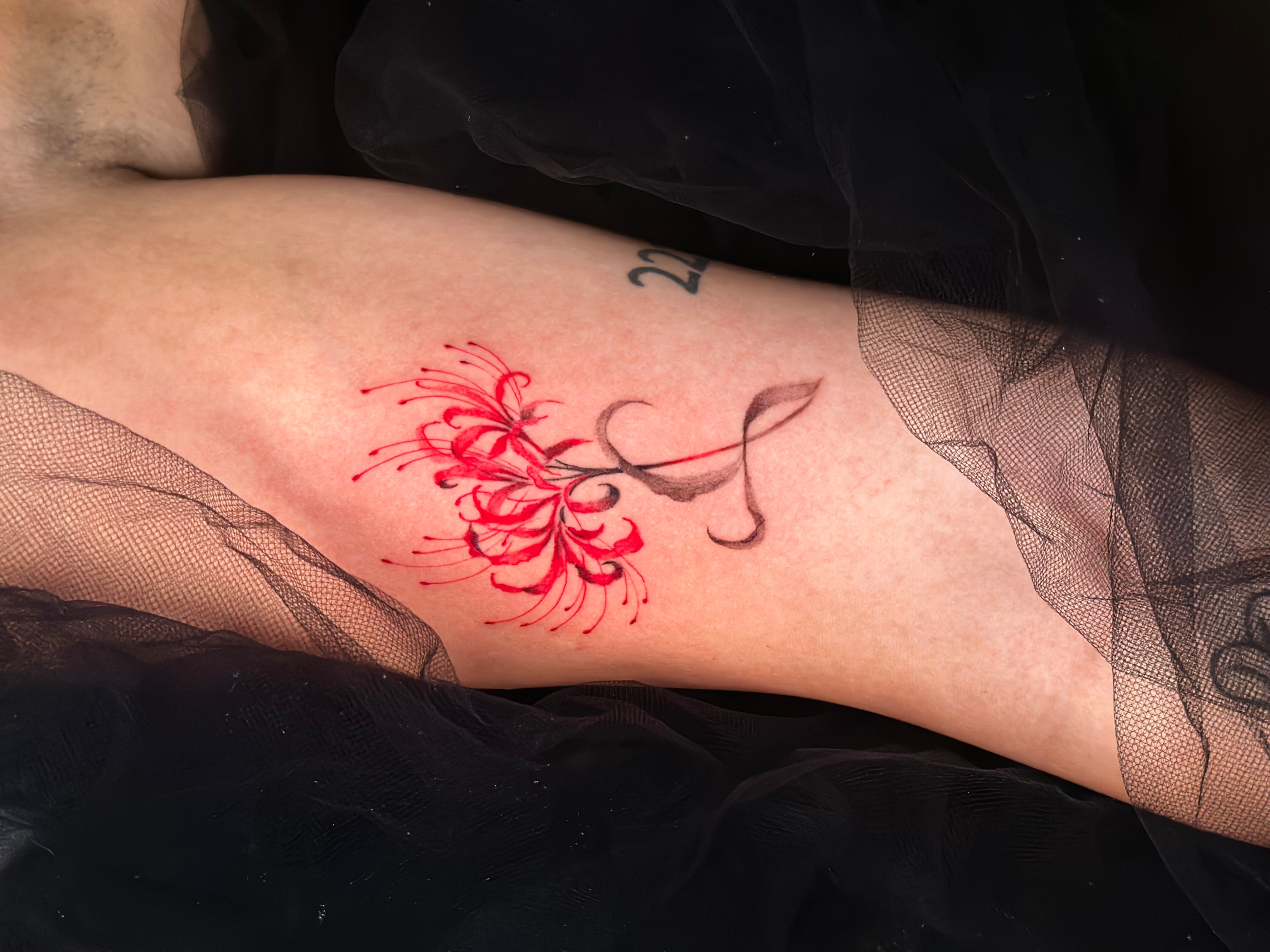Tattoo of a red flower with a black stem and leaves on a person's arm, surrounded by black fabric.