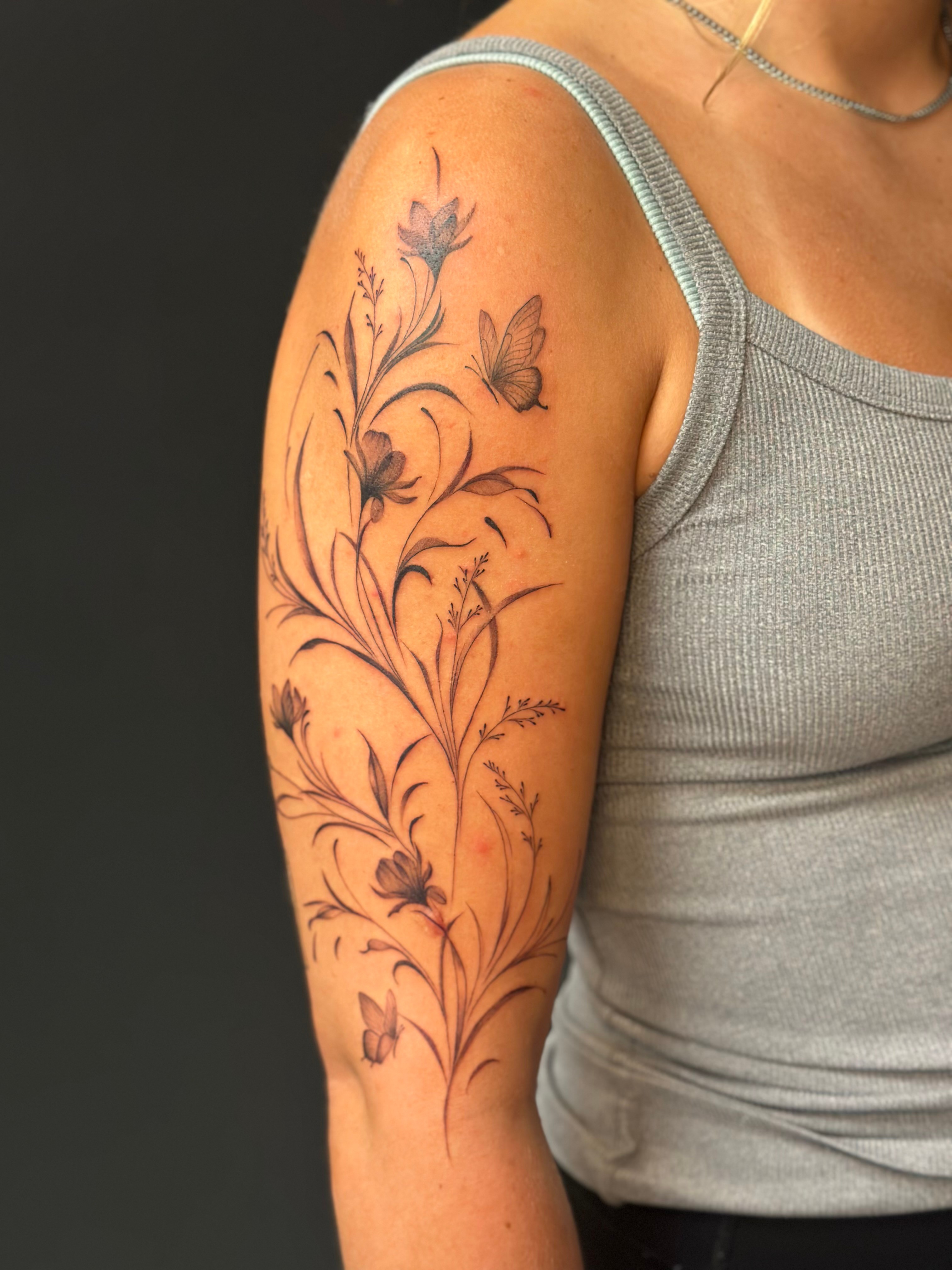 Tattoo of flowers and butterflies on person's upper arm and shoulder, wearing a gray tank top, against a dark background.