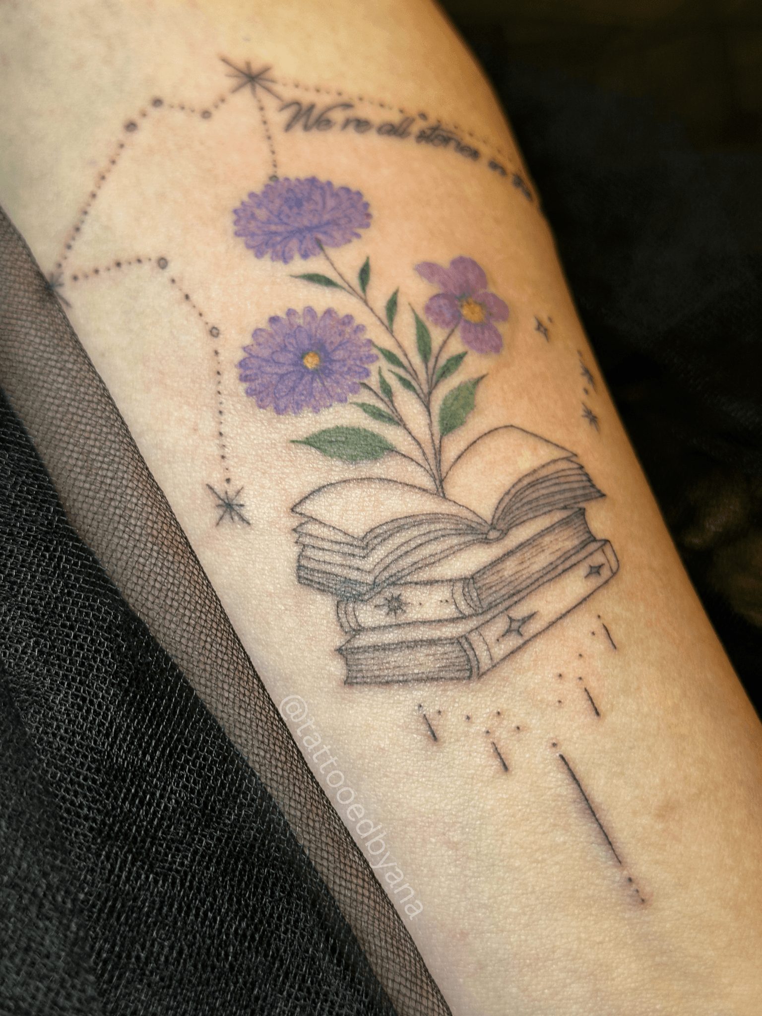 Tattoo of flowers emerging from open books, with constellation and handwritten text above, on skin.
