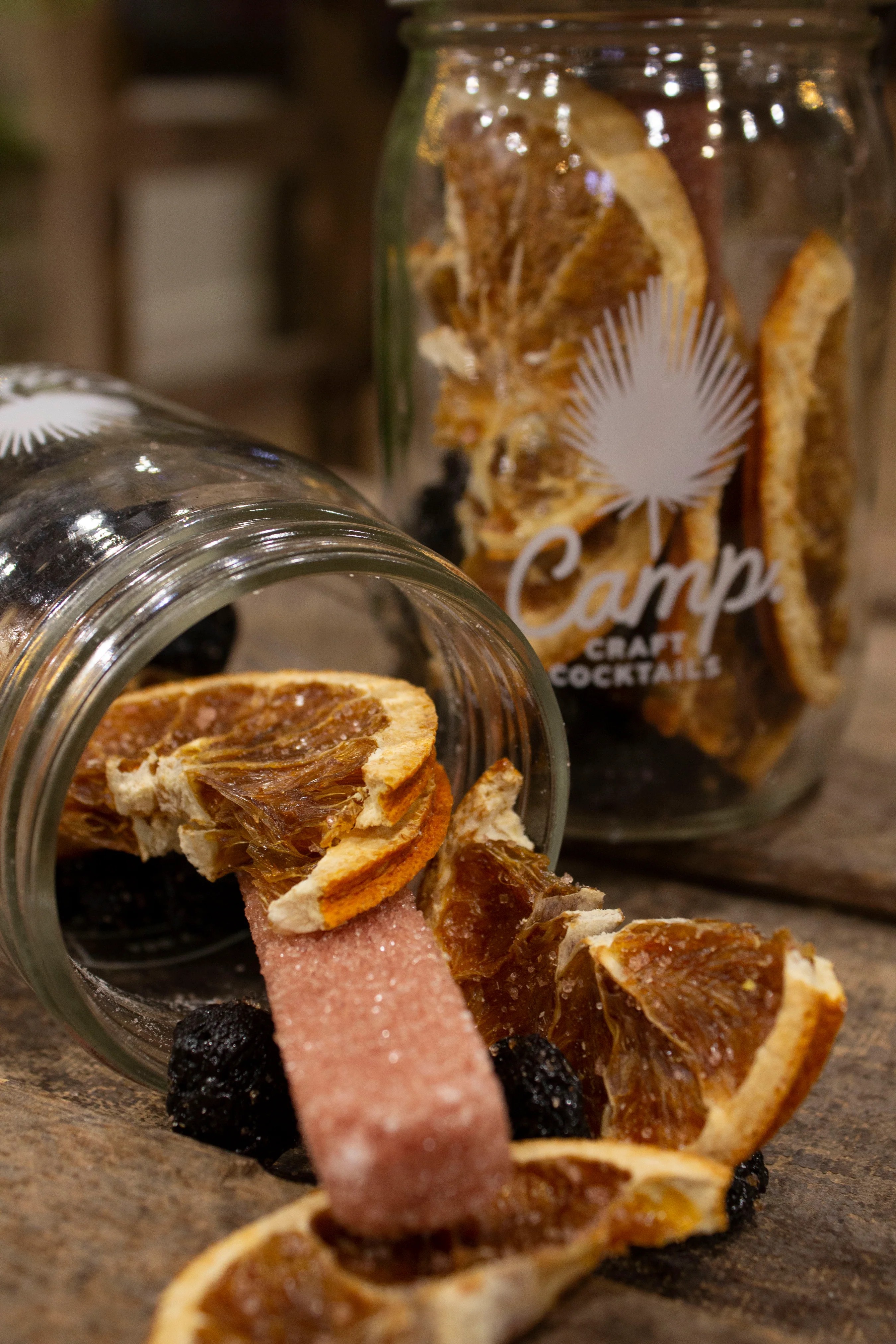 Jar with dried orange slices and a pink candy, some orange slices and candy outside the jar, on a wooden surface.