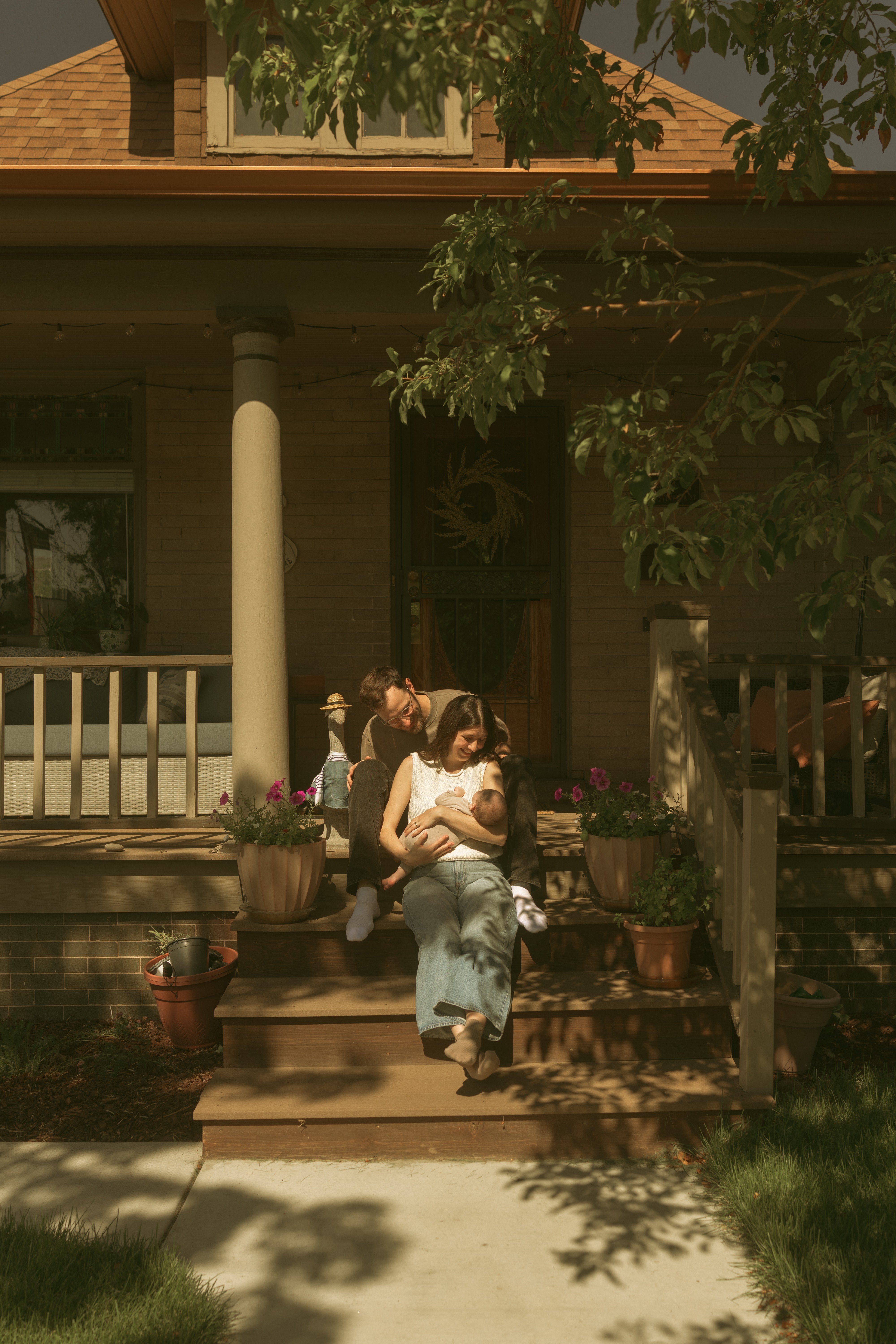 Two women sit on front steps of a house, one holding a child, surrounded by potted plants, with a porch and trees nearby.