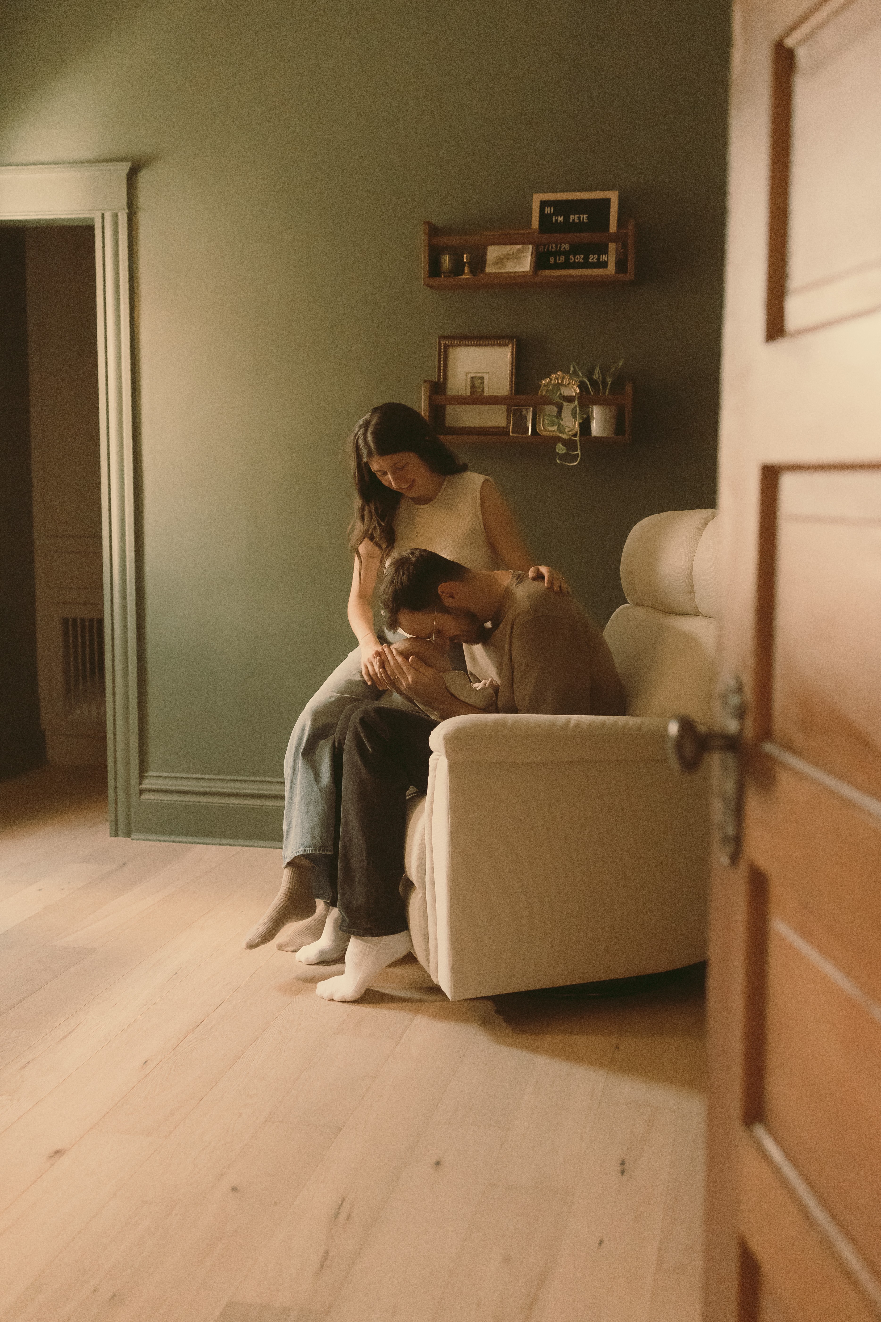 Two women sit on a white armchair in a room with green walls, one standing and the other sitting, with shelves on the wall behind them.