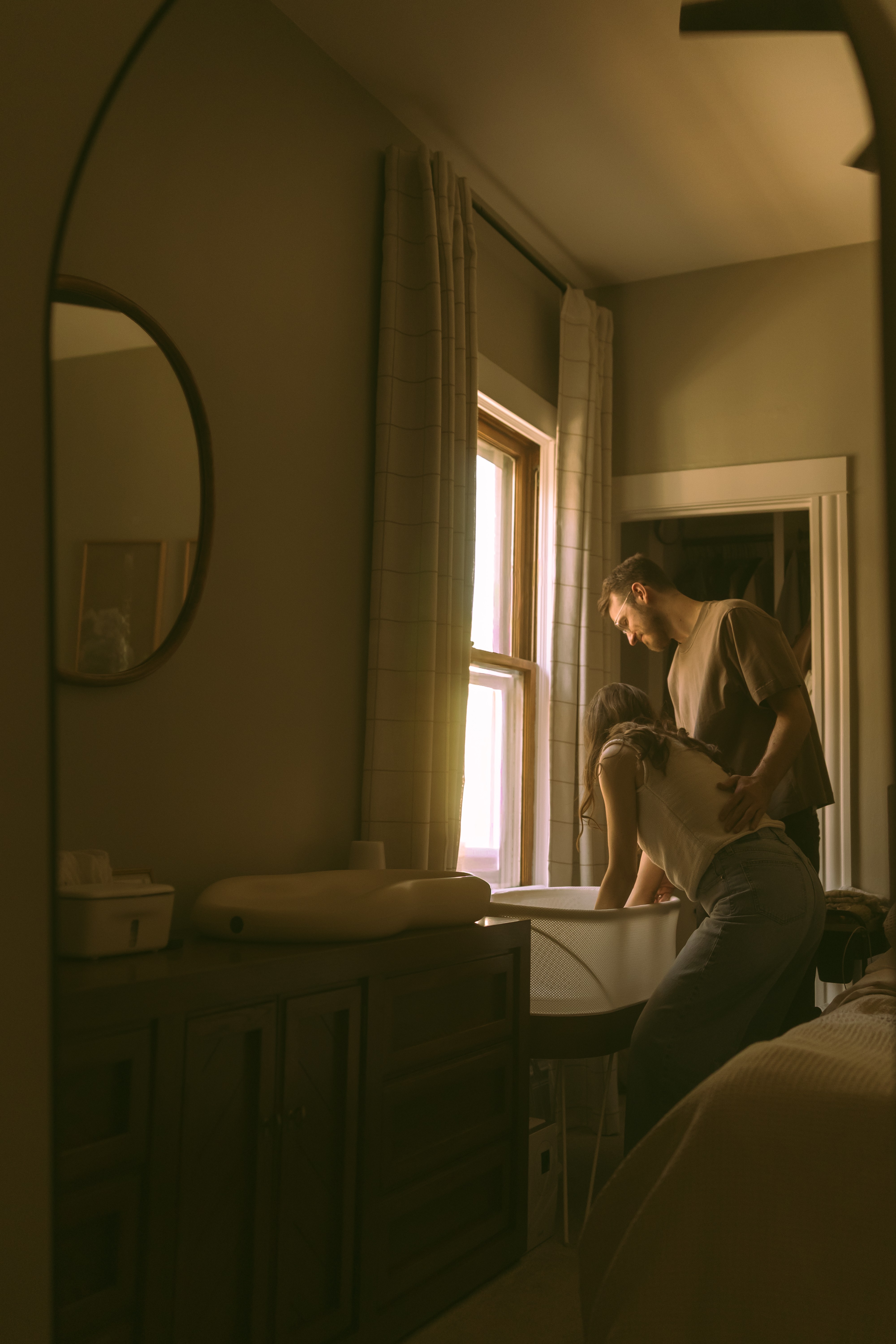 Two people by a window, one leaning on a table, in a dimly lit room with curtains and a mirror.