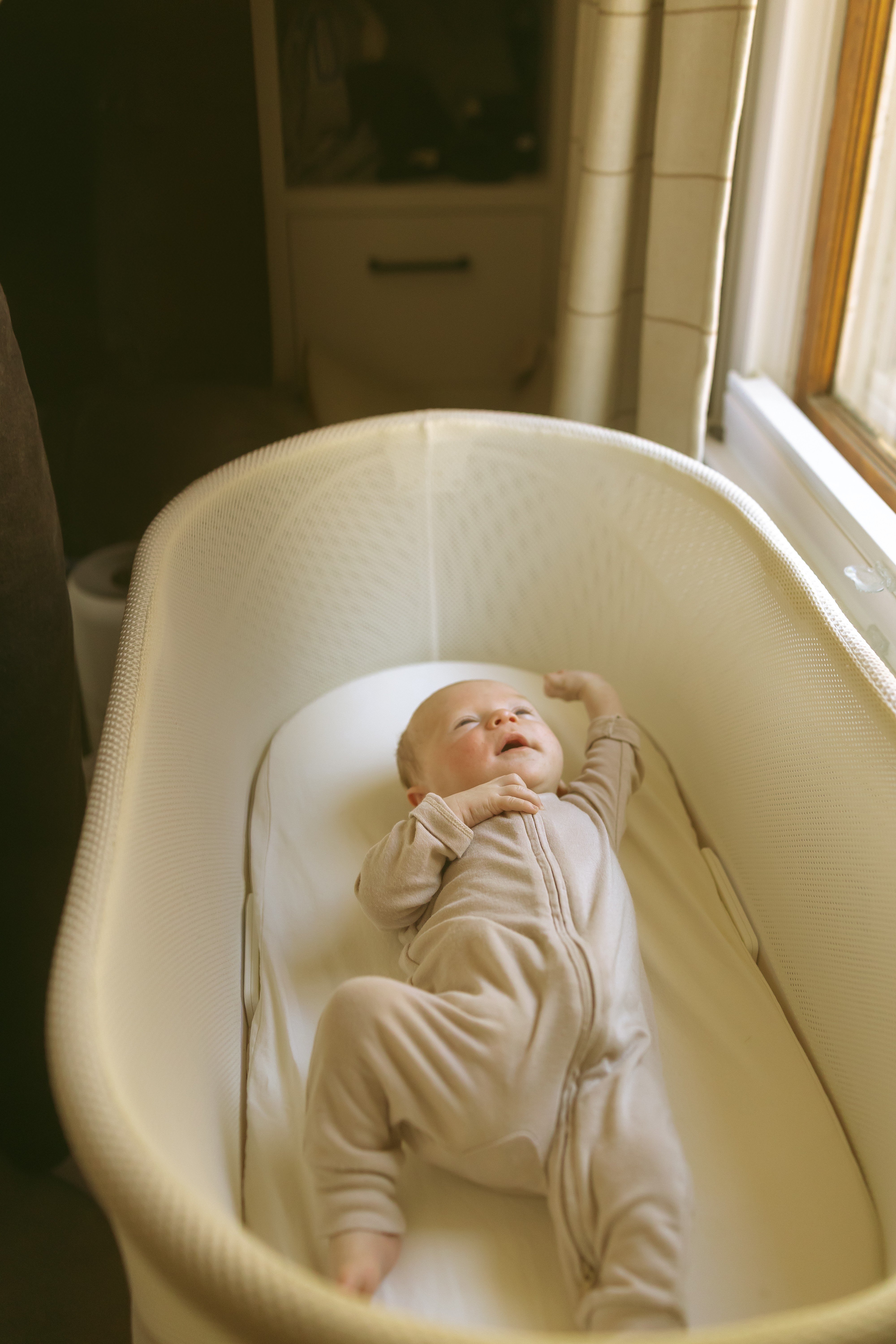 Baby lying in a bassinet near a window, looking up, with sunlight coming in.