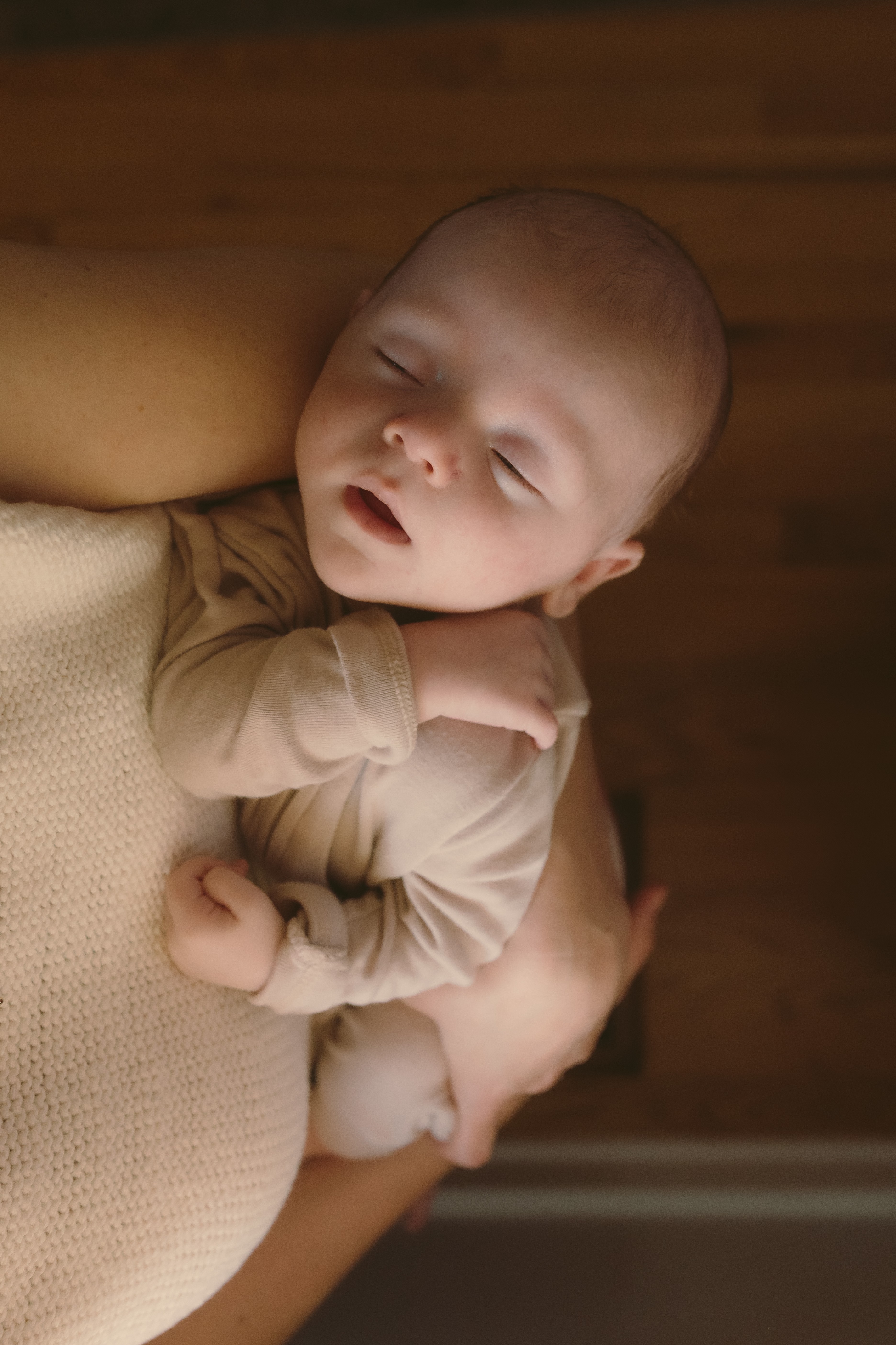 Baby sleeping peacefully, resting on an adult's arm, with a wooden background behind them.