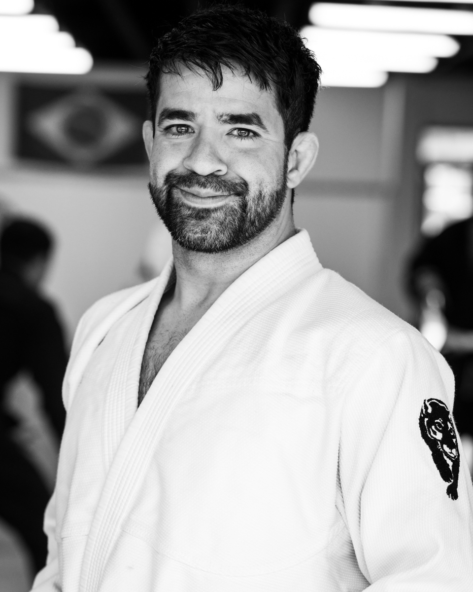 Man with dark hair and beard smiling, wearing a martial arts gi, in a training facility, black and white photo.