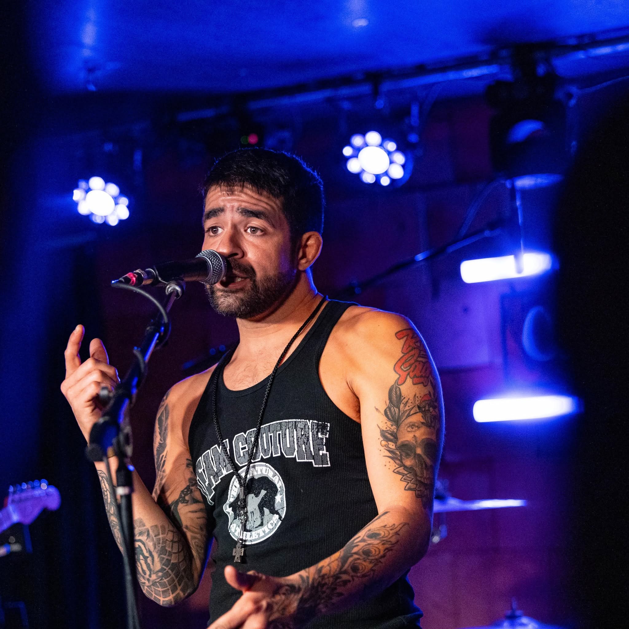 Male singer with tattoos performs on stage with blue lighting, wearing a black tank top, holding a microphone.