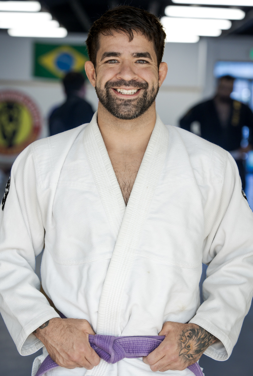 Smiling man in a white martial arts uniform with a purple belt, standing indoors with people in the background.