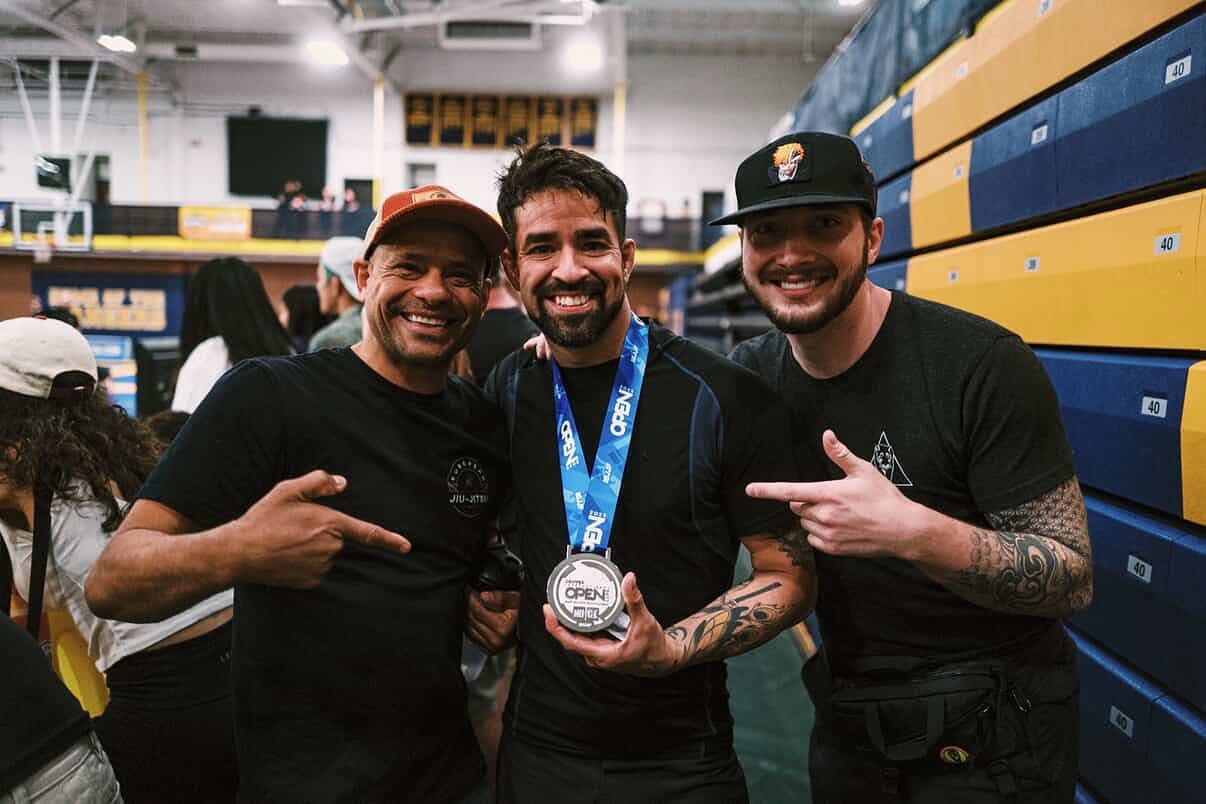 Three smiling men in a gymnasium, one holding a medal, with people and gym equipment in the background.