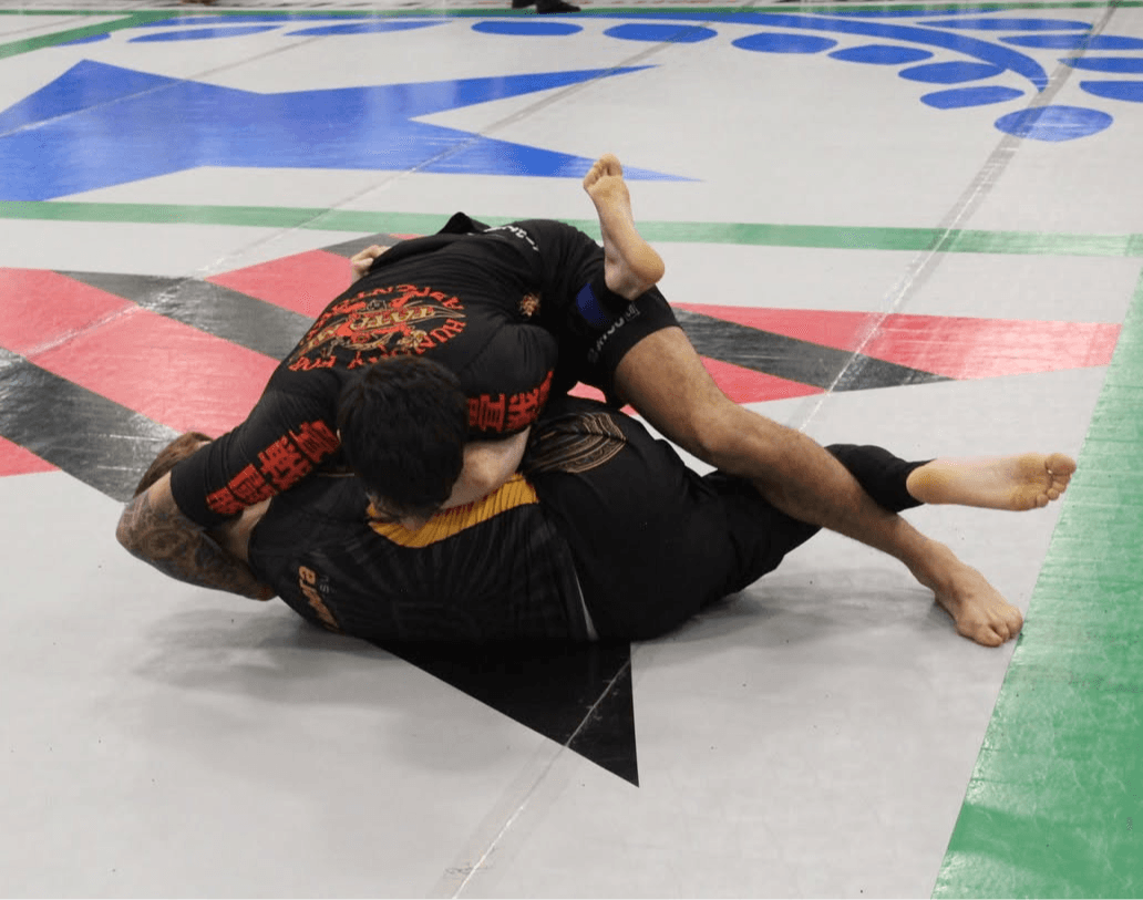 Two people grappling on a mat, one on top of the other, in a Brazilian Jiu-Jitsu match.