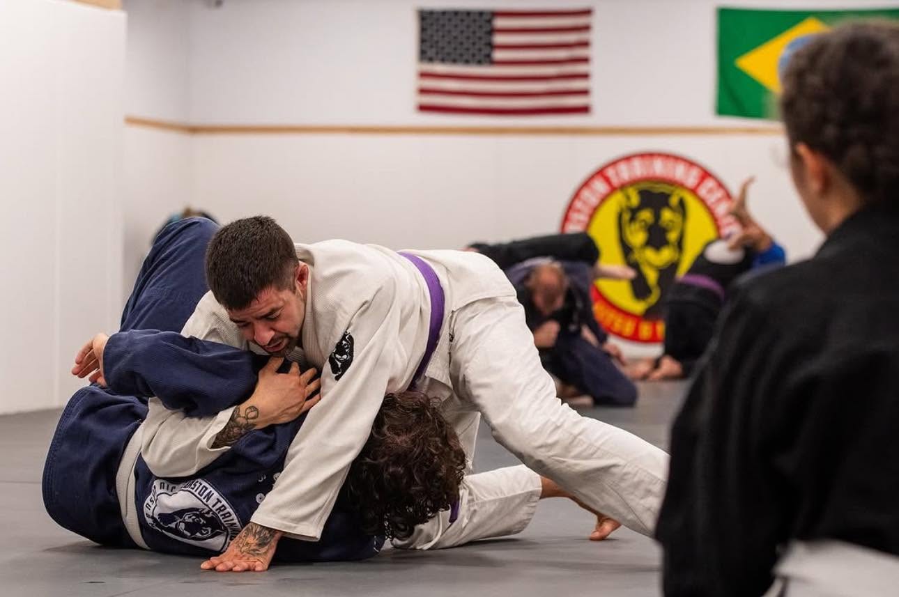 Two people practicing Brazilian Jiu-Jitsu on mats, with flags and a logo in the background, one person on top of the other.