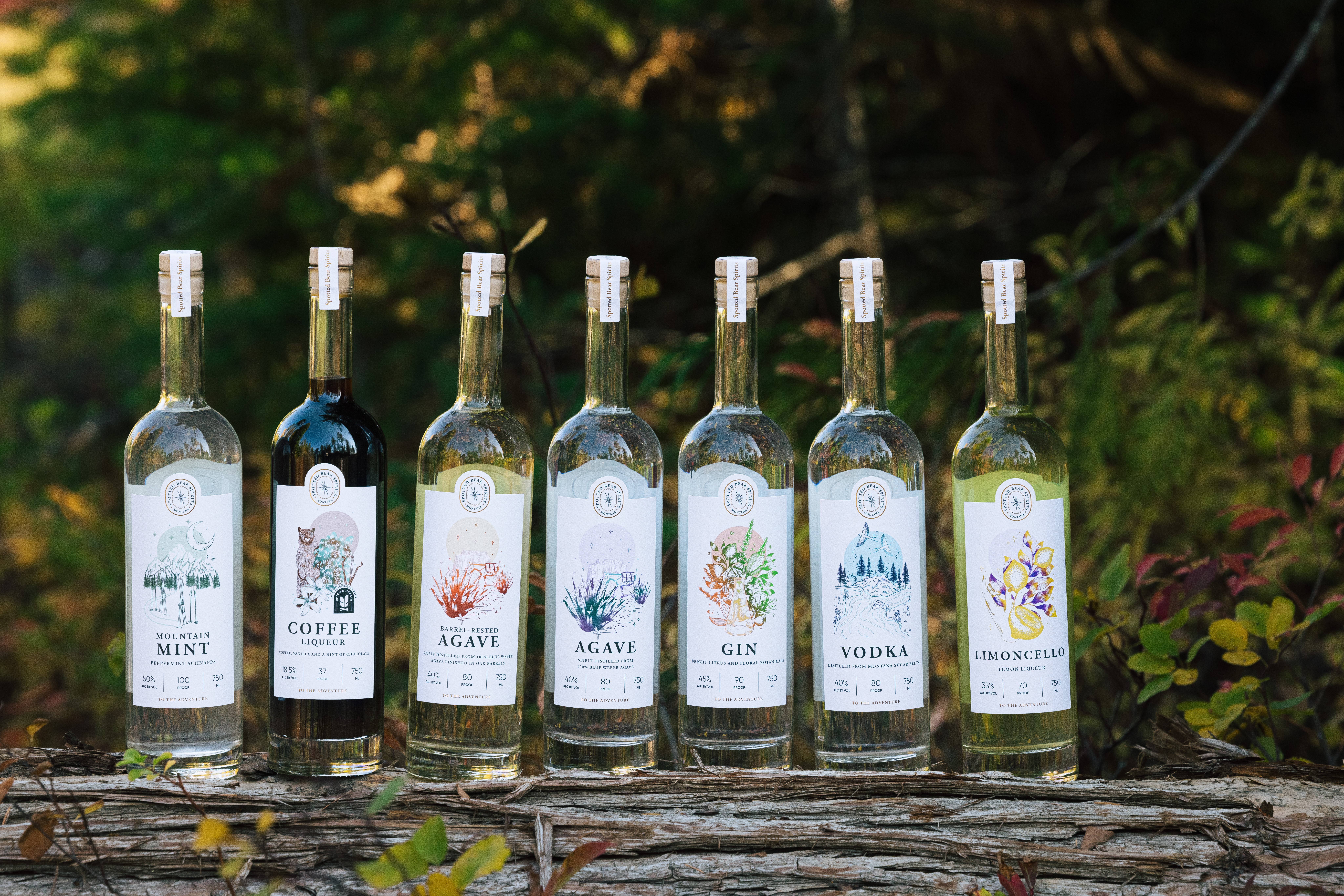 Six bottles of flavored spirits lined up on a log outdoors with greenery in the background.