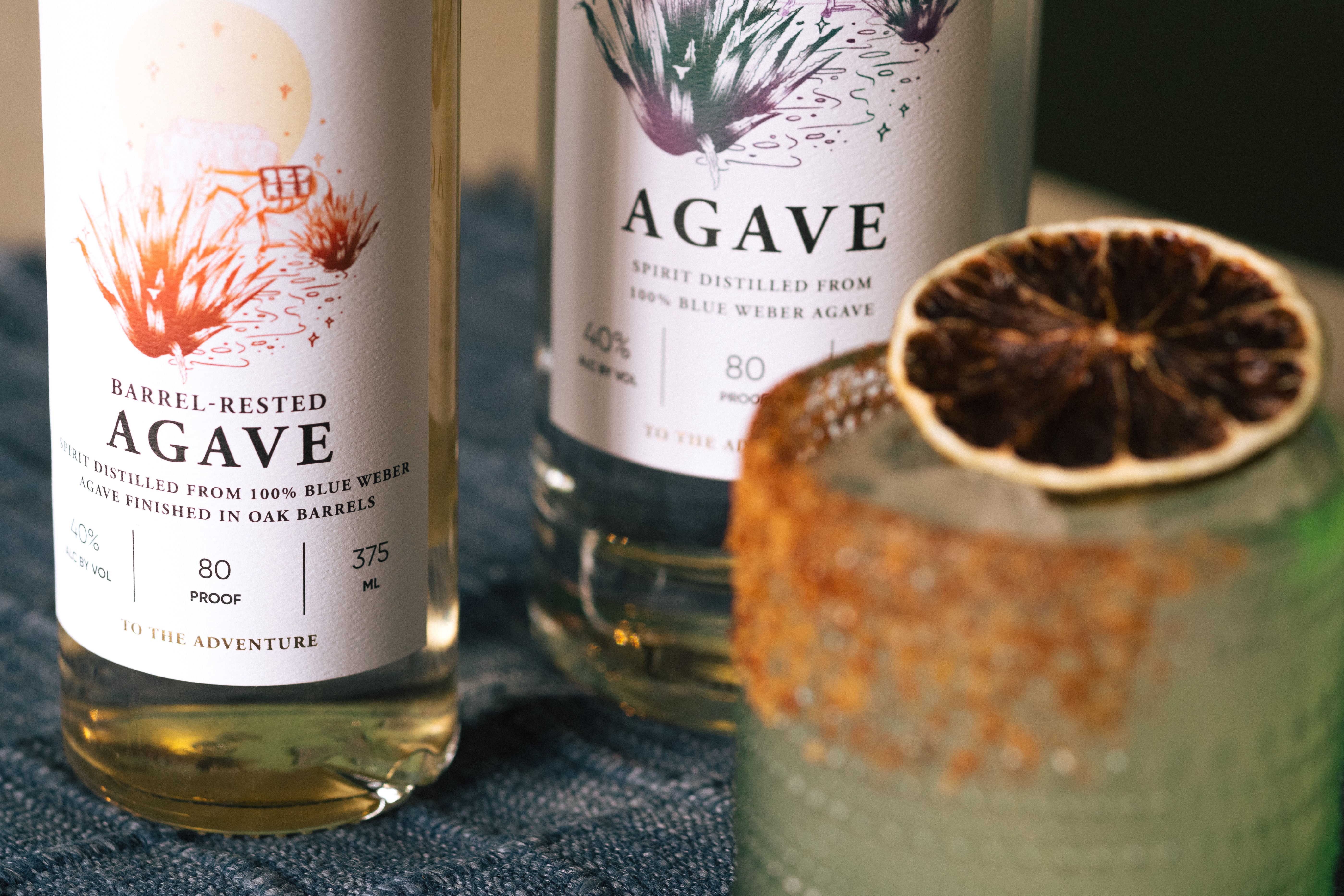 Two bottles of agave spirit and a glass with a dried citrus slice garnish, on a dark surface.