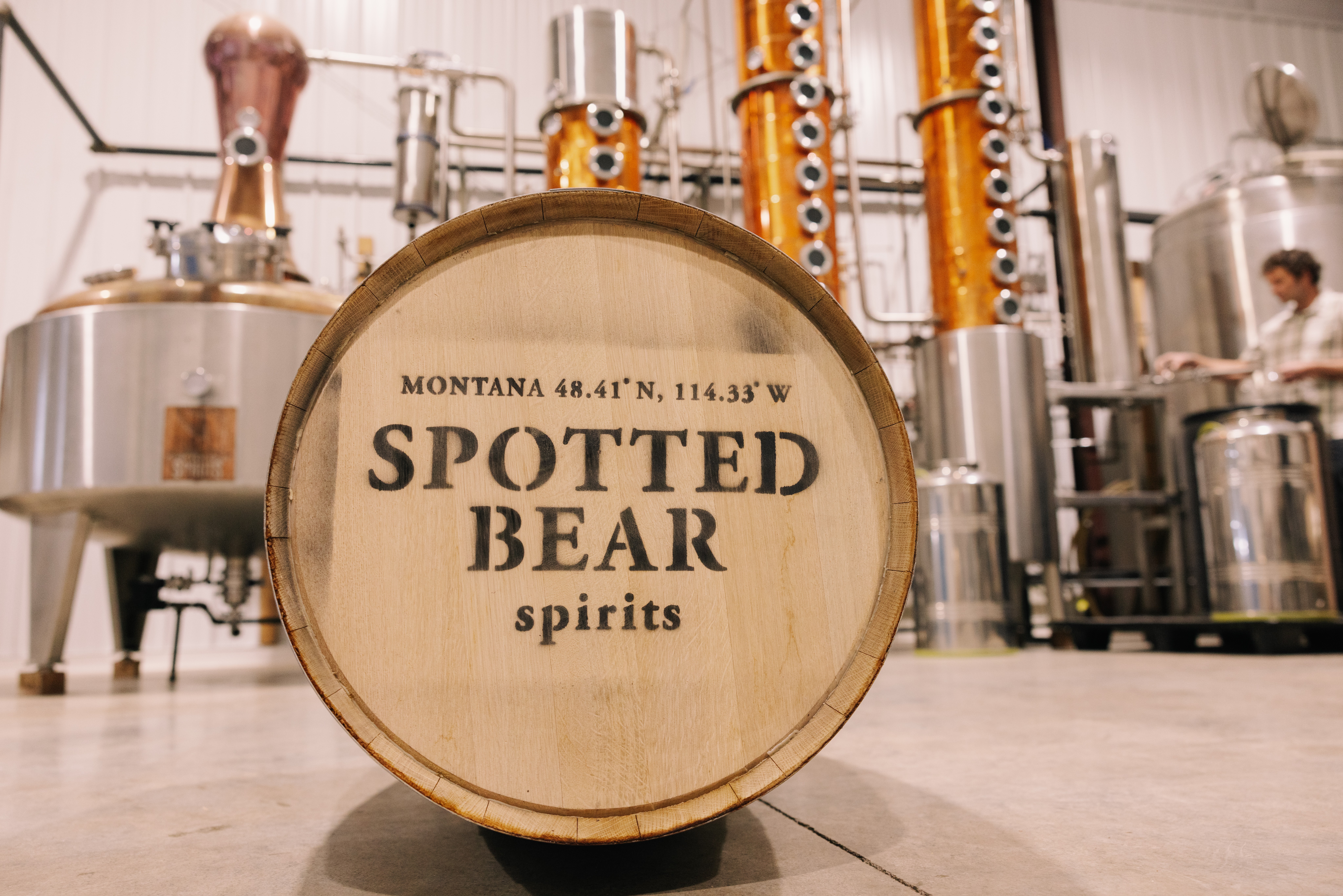 Barrel with text 'Spotted Bear spirits' in a distillery with equipment in background.