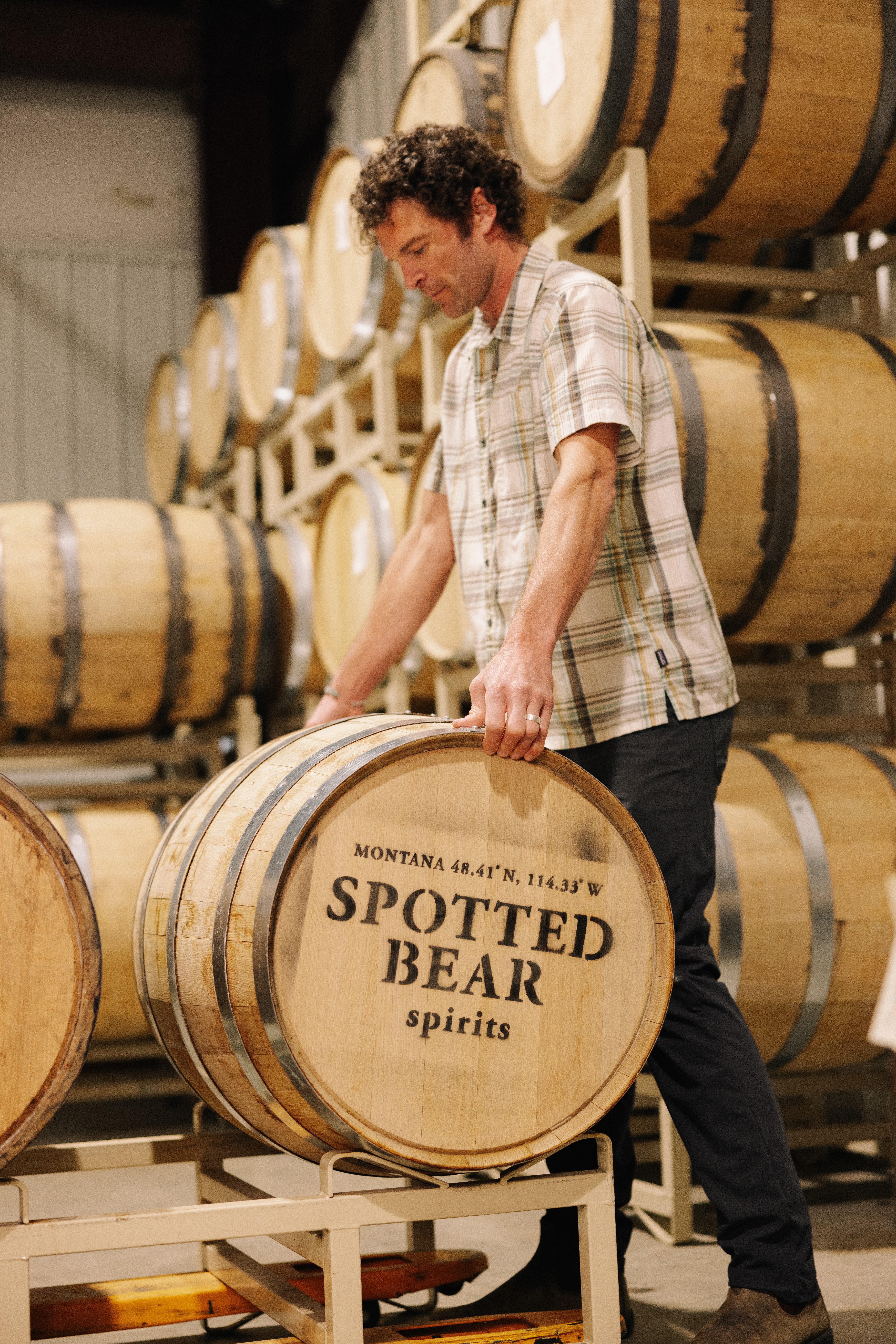 Man in plaid shirt standing next to a barrel labeled 'Spotted Bear spirits' in a warehouse with stacked barrels