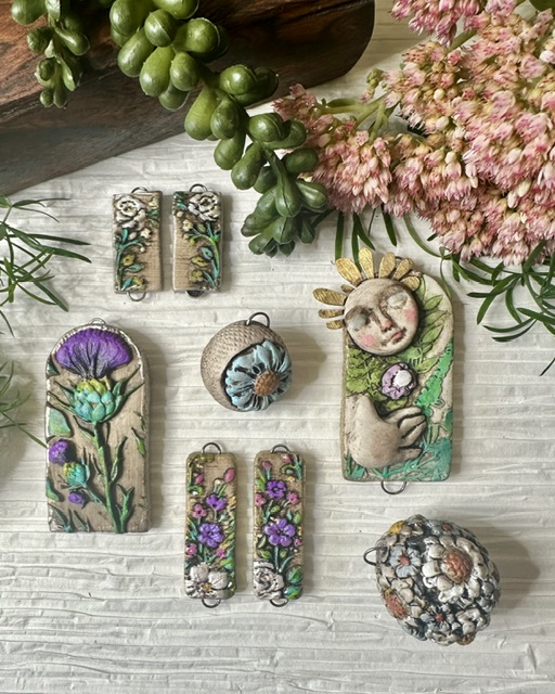 Jewelry pieces with floral and nature designs arranged on a white wooden surface, surrounded by pink flowers and green foliage.