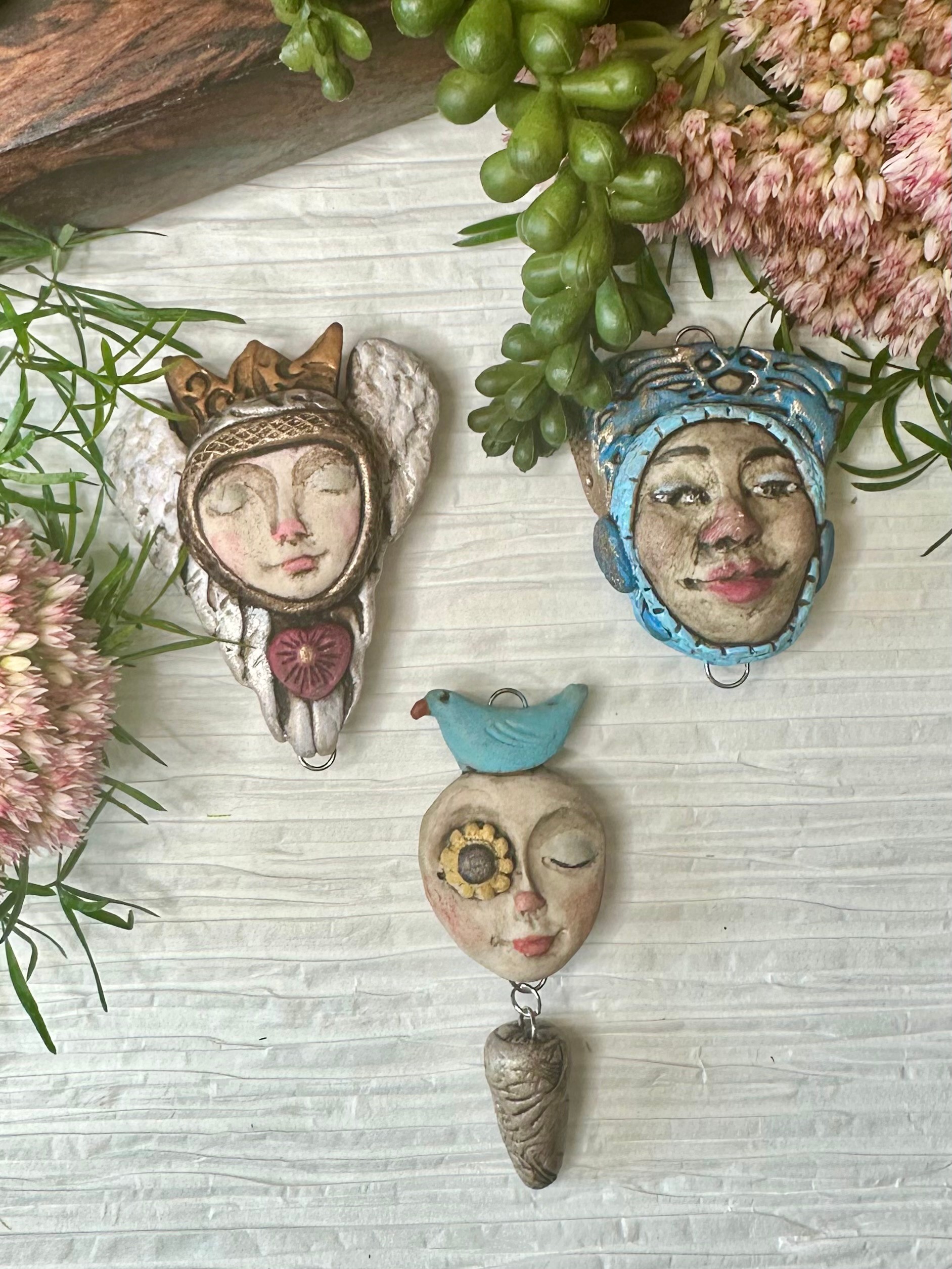 Four decorative pins with faces, surrounded by pink flowers and green plants, on a white wooden surface.