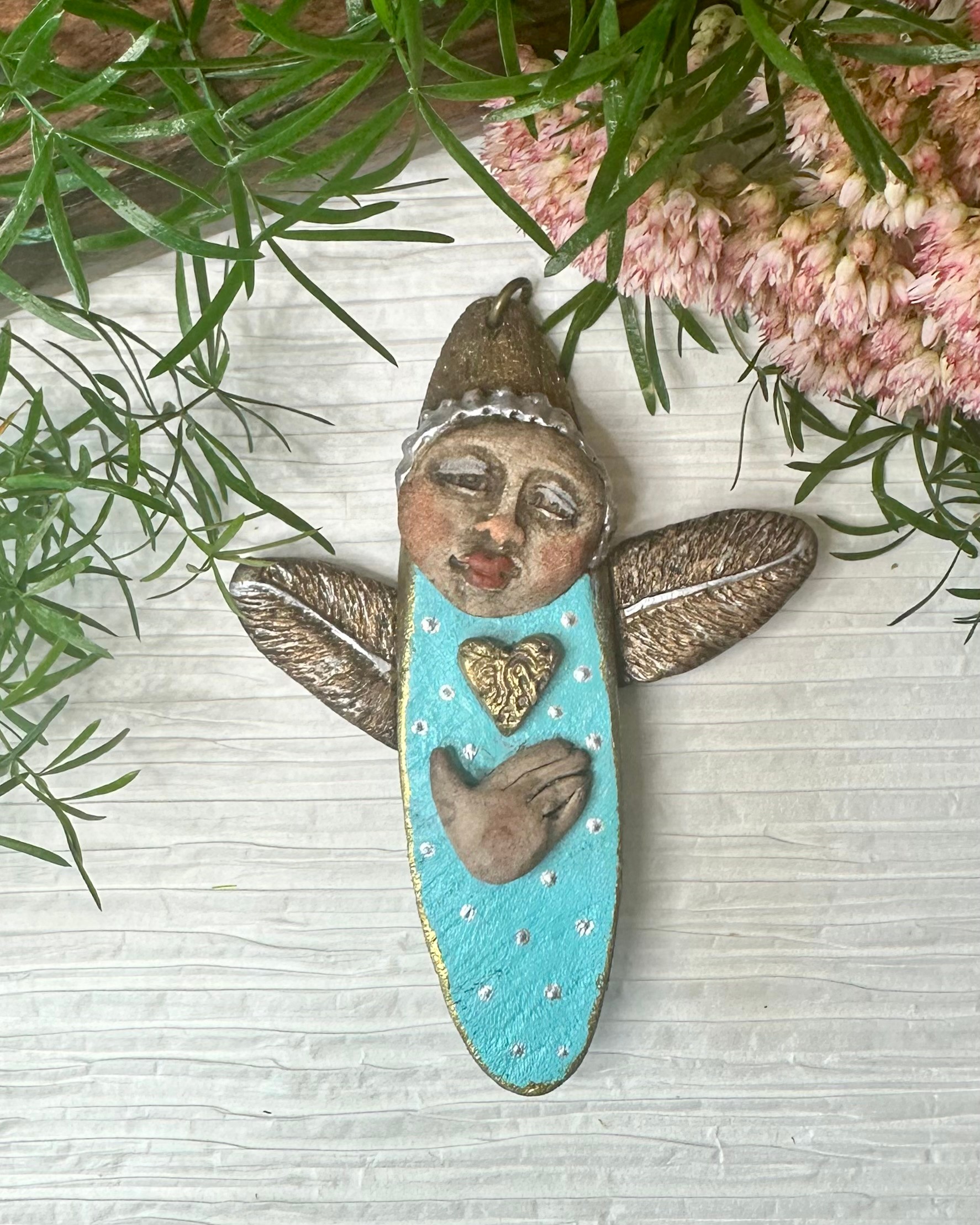 Decorative butterfly-shaped ornament with a painted face, blue body, and heart-shaped cutouts, surrounded by pink flowers and green leaves.