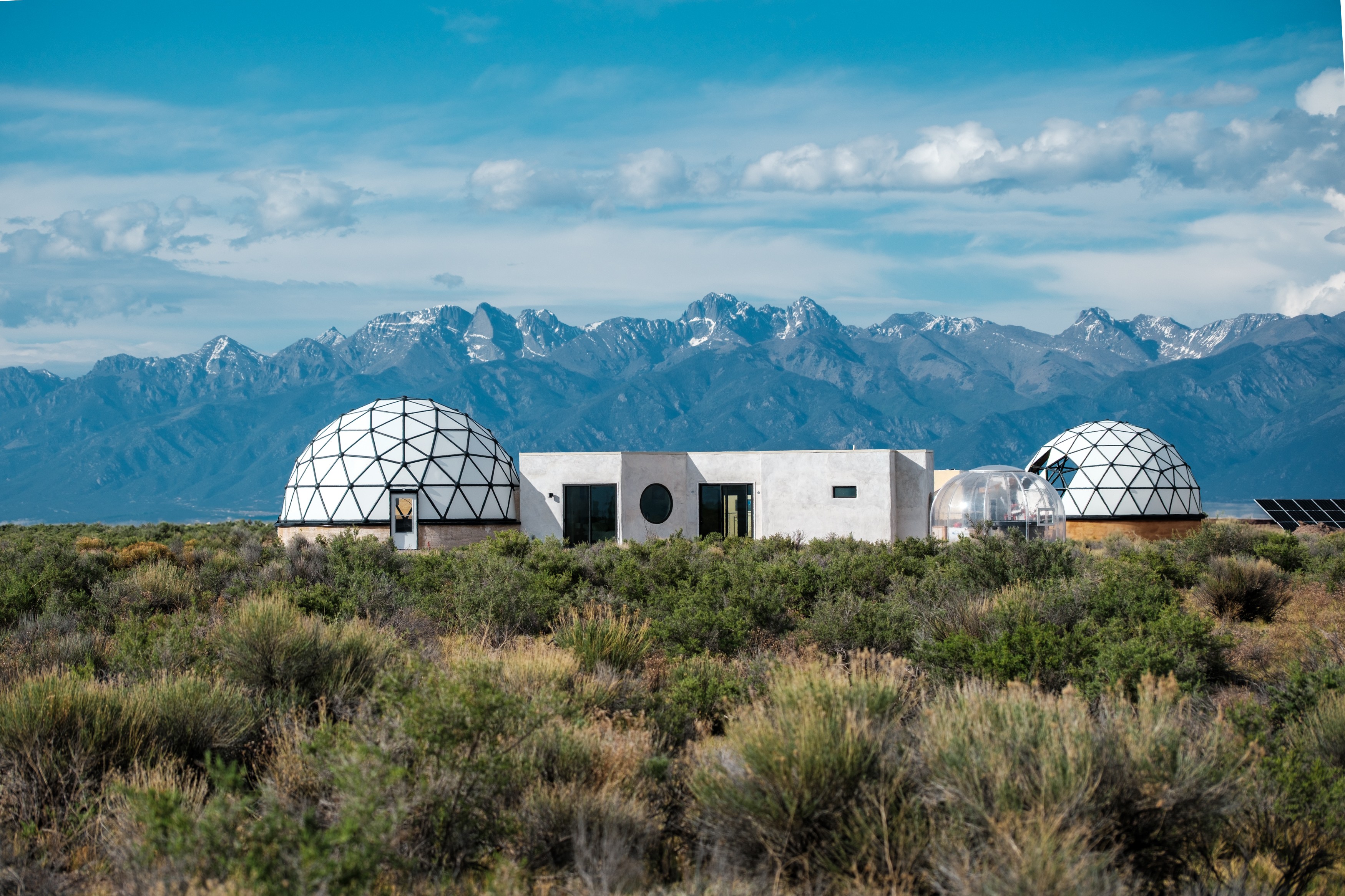 Two geodesic domes and a rectangular building in a desert landscape with mountains in the background, under a partly cloudy sky.