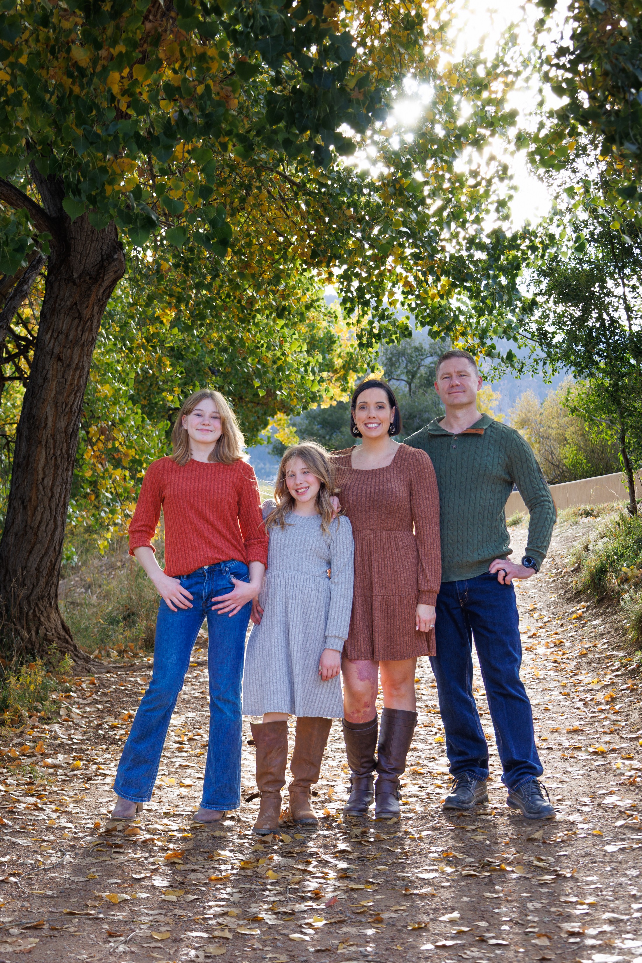 Four people standing outdoors on a leaf-covered path under a large tree, smiling, with sunlight filtering through leaves.