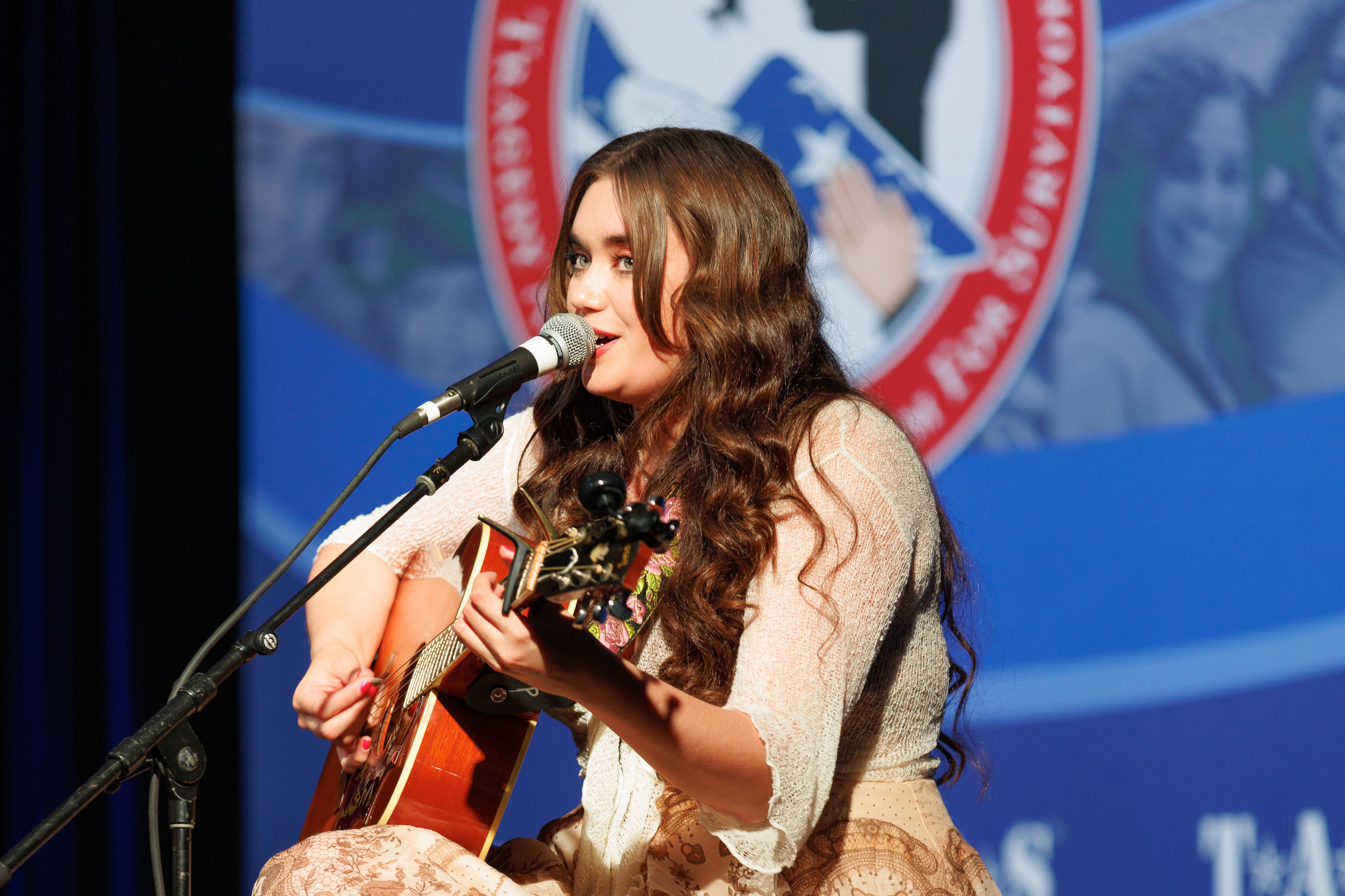 Young woman with long wavy hair singing into a microphone while playing an acoustic guitar on stage.