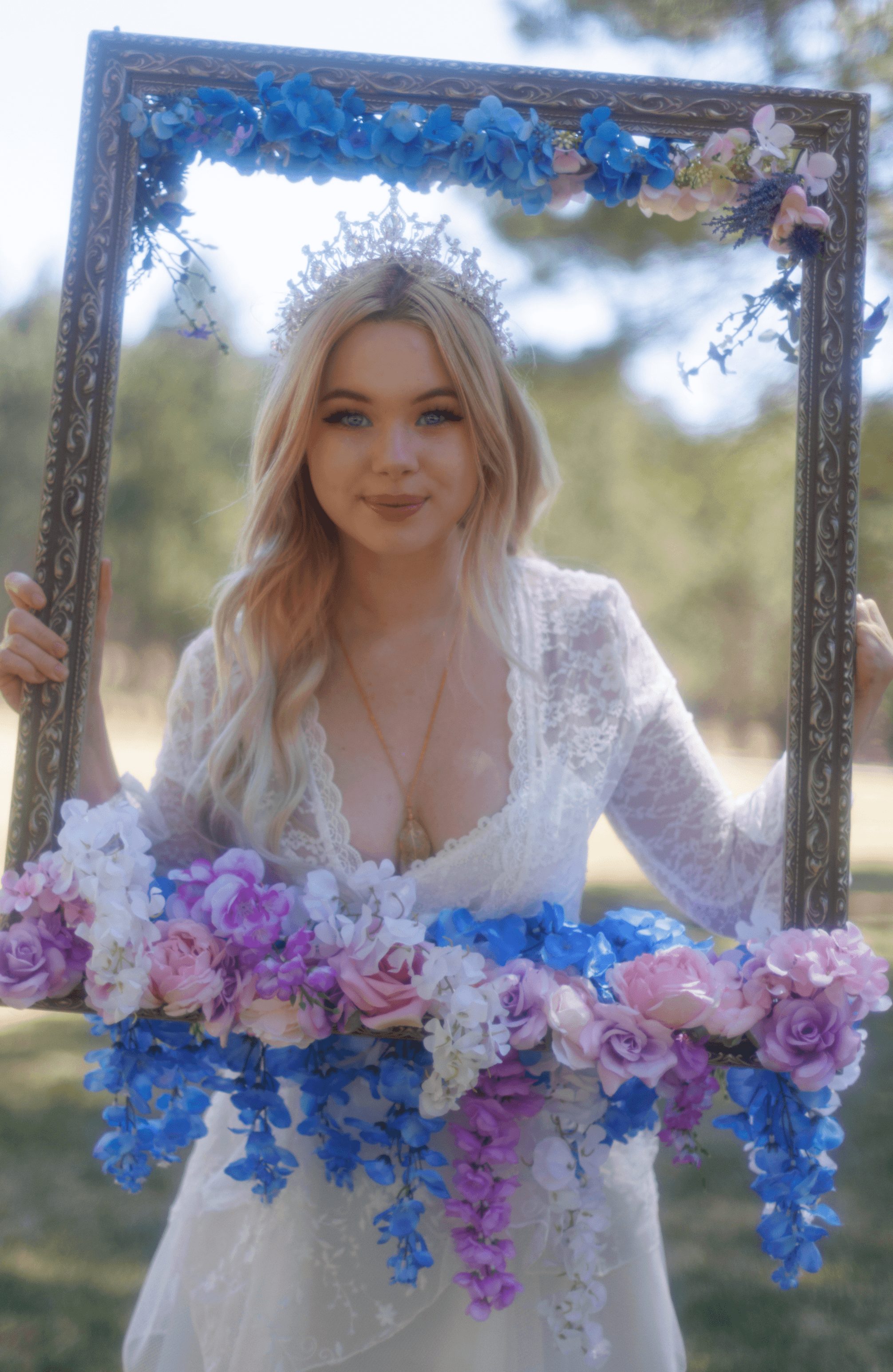 Young woman with blonde hair in a white dress holding a floral frame outdoors, surrounded by flowers and greenery.