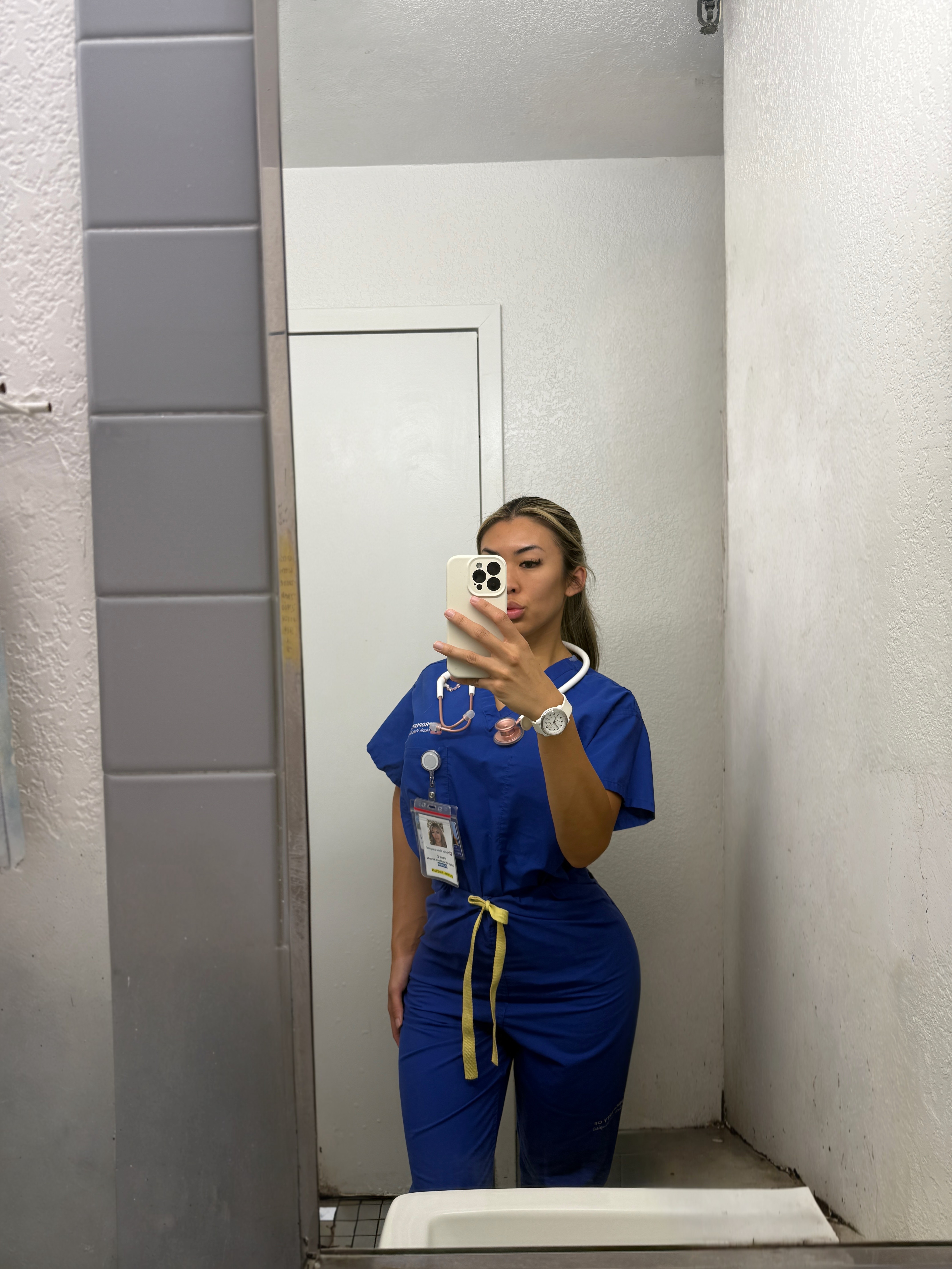 Young woman in blue medical scrubs taking a mirror selfie in a small room, holding a phone with a white case.