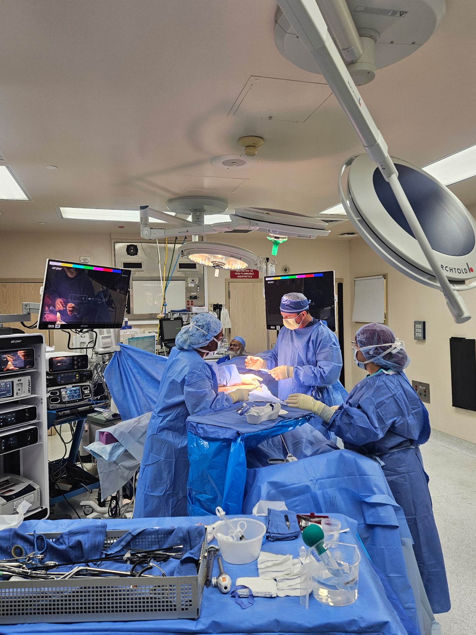 Medical team performs surgery in an operating room with monitors and equipment, all wearing blue scrubs and surgical masks.