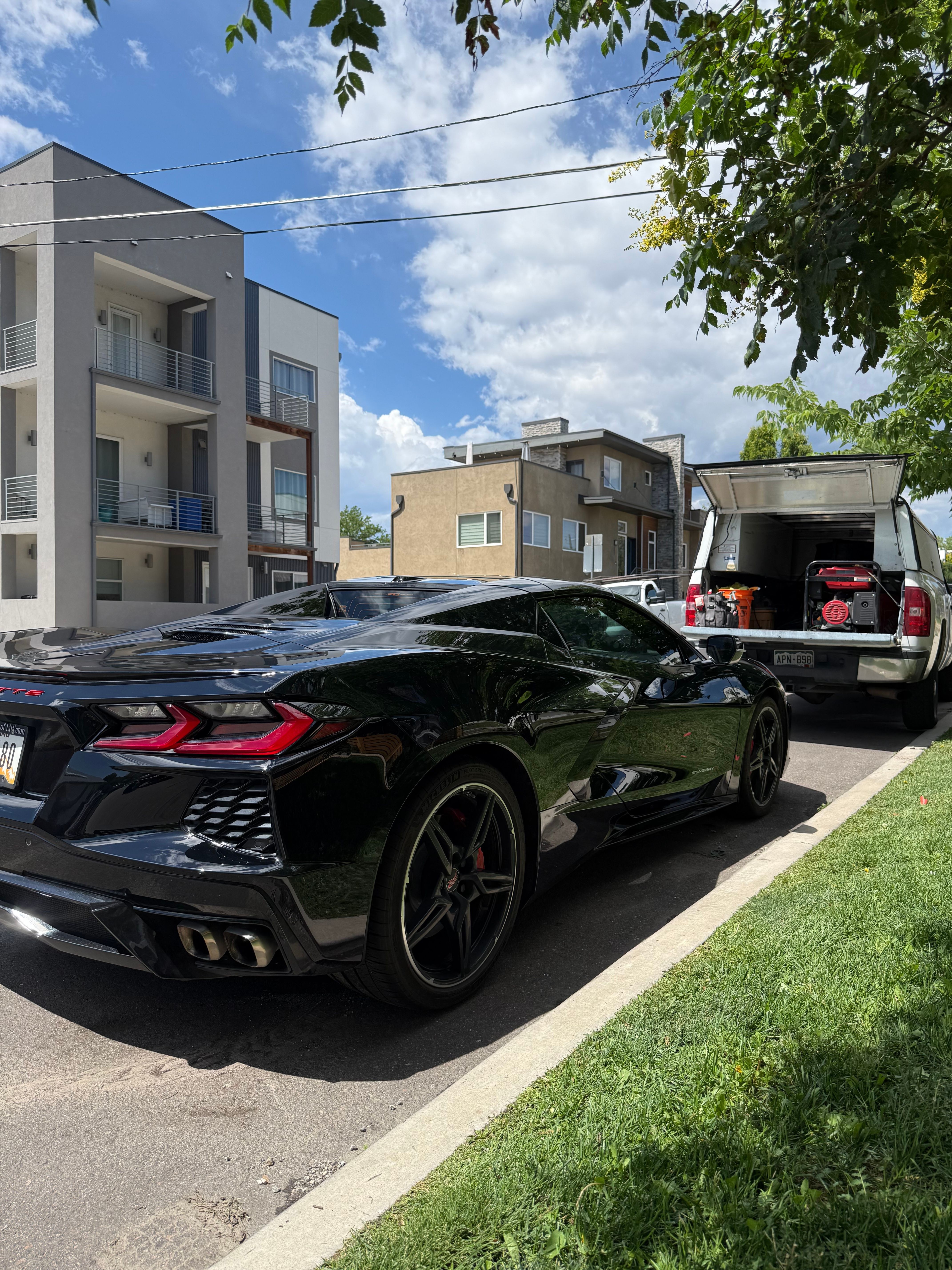 Black sports car parked on a residential street with buildings, trees, and a blue sky with clouds.