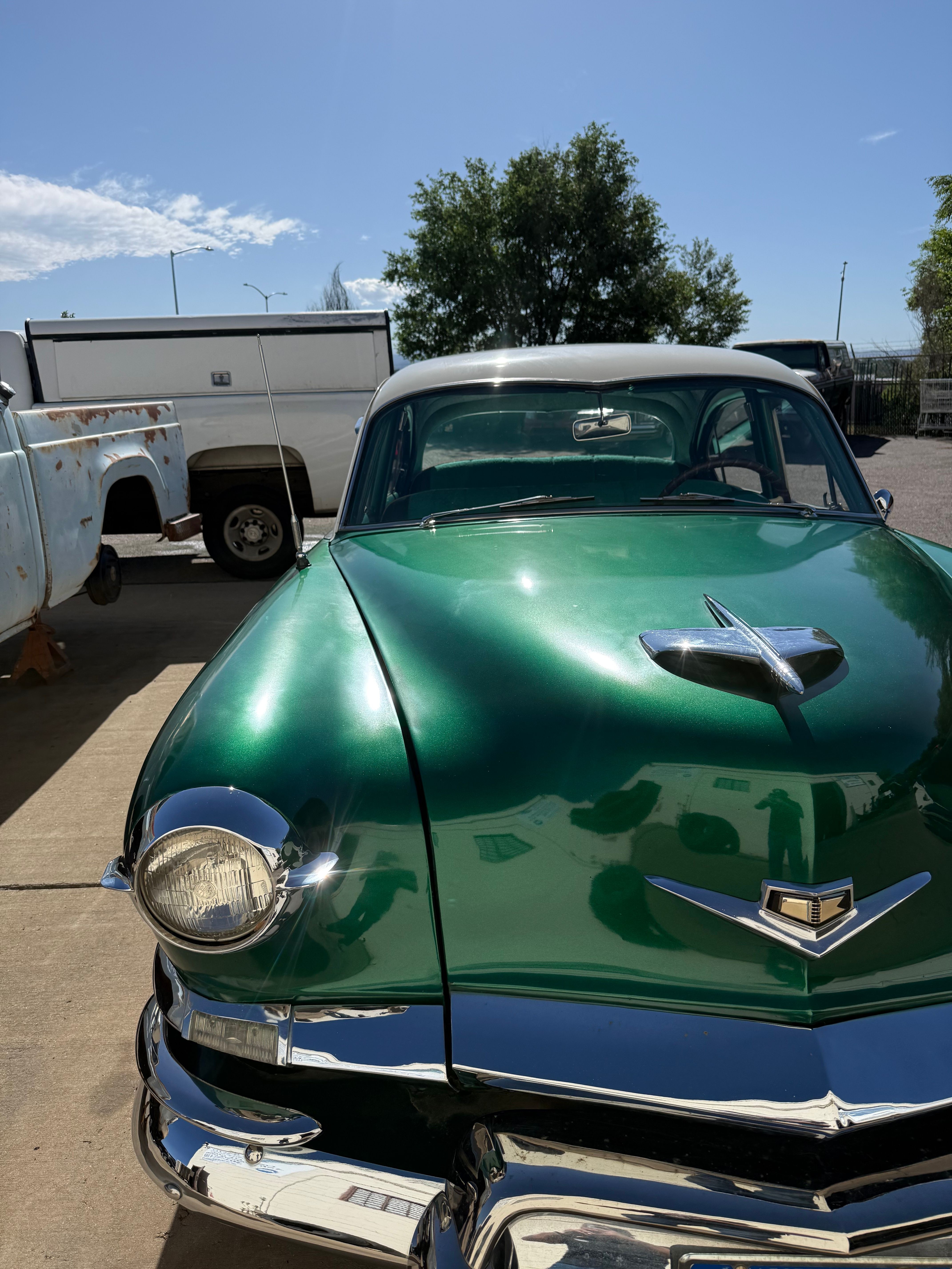 Green vintage car with chrome details parked outdoors, with trees and a trailer in the background.
