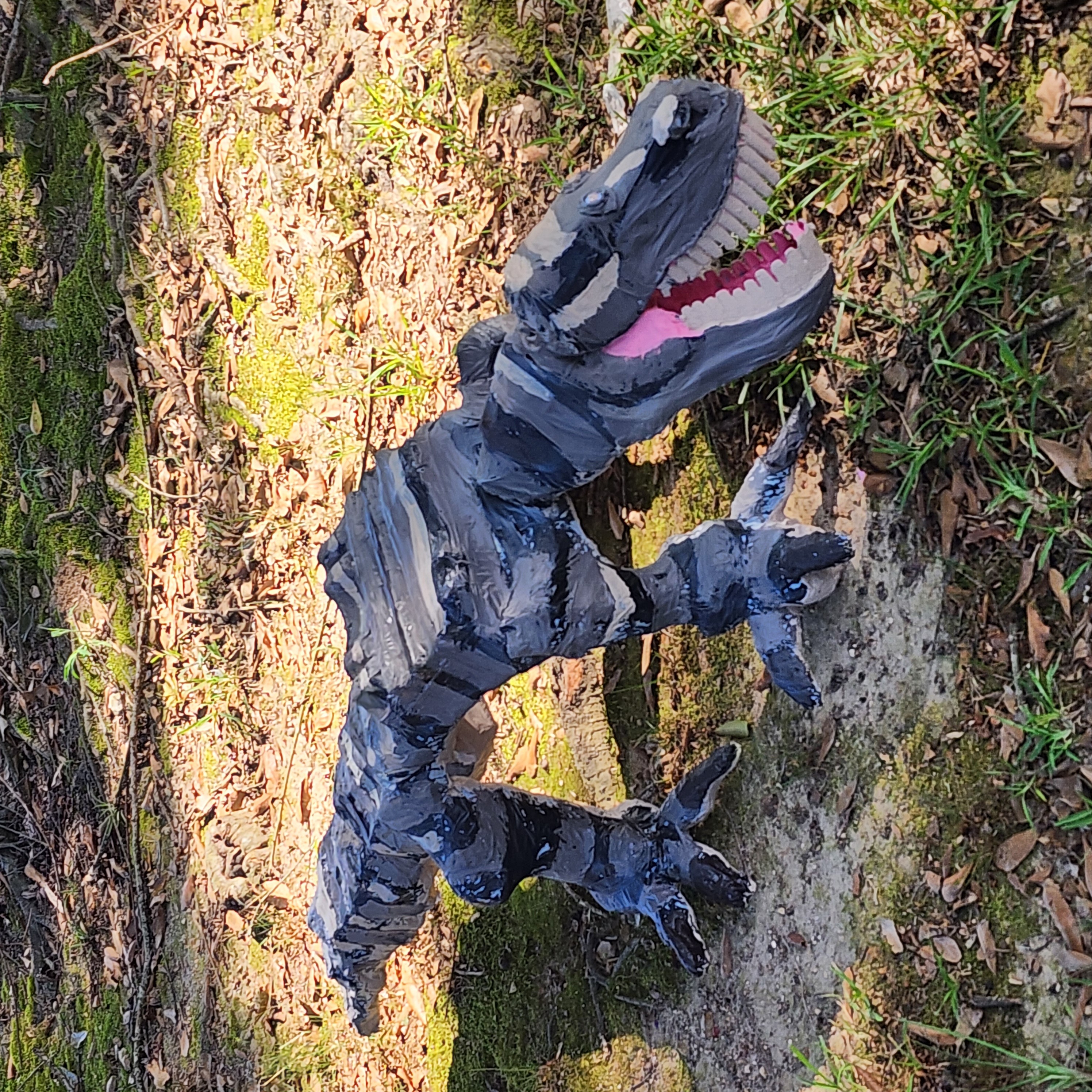 Dinosaur model with open mouth and sharp teeth, standing outdoors on grass and dirt, facing right.