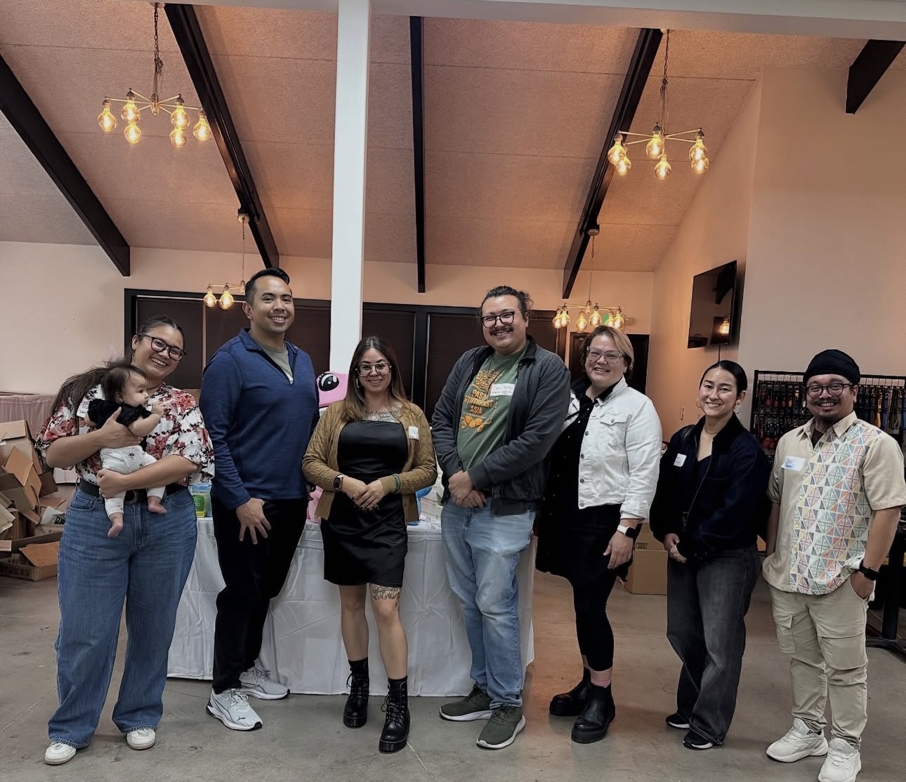 Group of seven people standing indoors, smiling, with a high ceiling and hanging lights, in a casual setting.