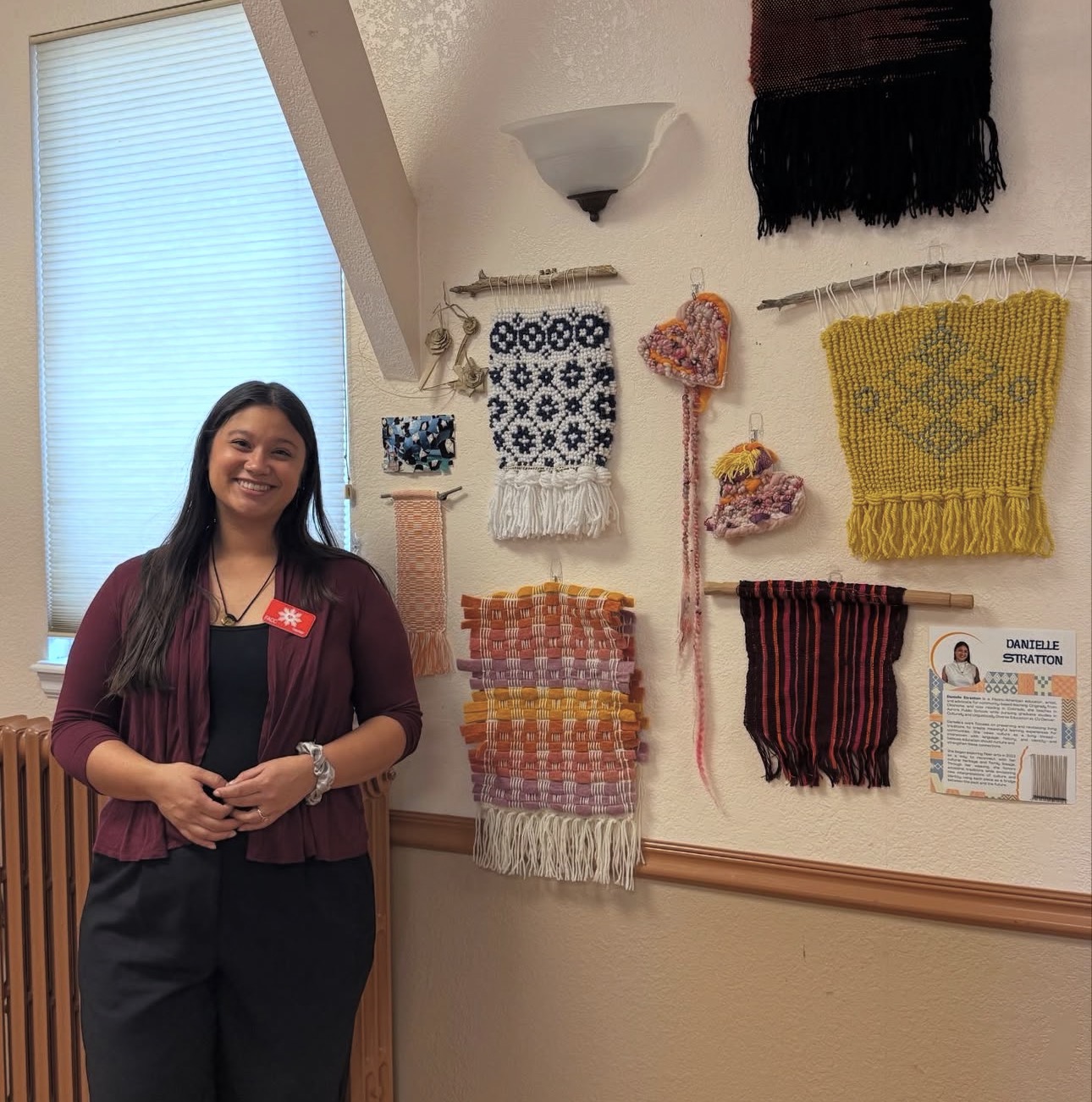 Woman standing indoors in front of a wall with colorful woven textile art pieces, smiling, wearing a maroon jacket and black pants.